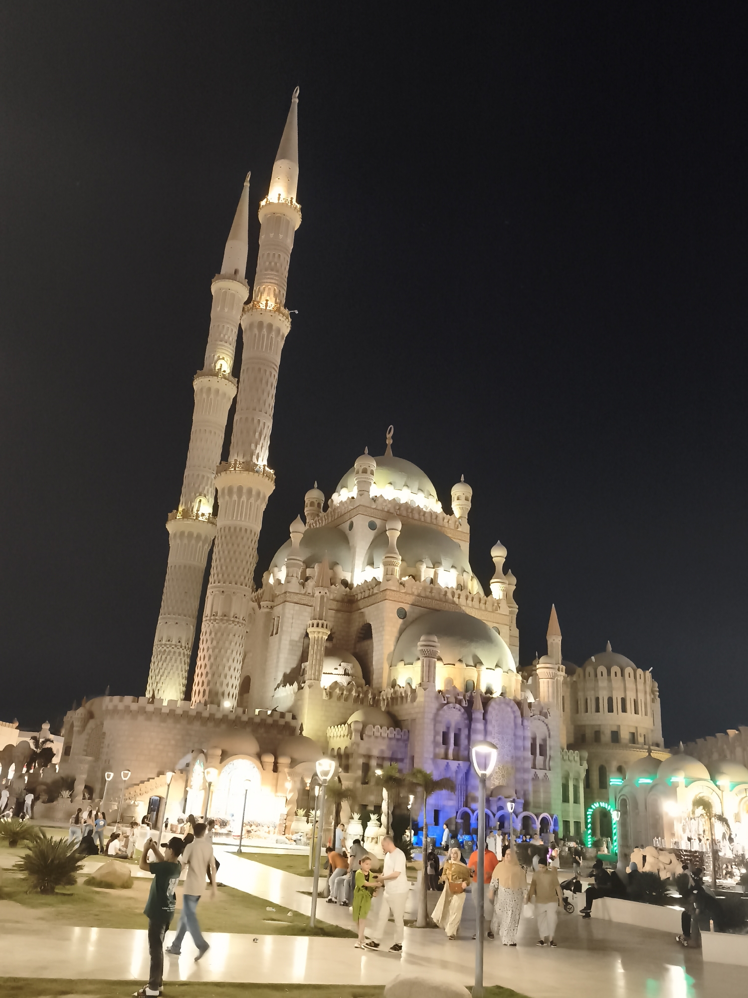 The image shows the Al Sahaba Mosque at night in Sharm El Sheikh, Egypt. The mosque, illuminated with warm lights, features distinctive architecture with multiple domes and tall minarets. A group of people are walking in the foreground, some taking pictures of the mosque. Palm trees and landscaping are visible around the mosque. The sky is dark, indicating it is nighttime.