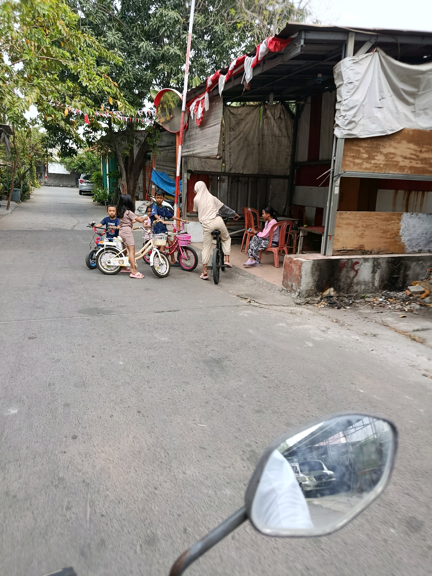 In Bekasi, Indonesia, an outdoor street scene depicts several children interacting with bicycles. A woman wearing a hijab and light-colored clothing is seated on a bicycle, appearing to be in motion. Nearby, two young children are near a beige bicycle, with one child standing on the pedal. Another child, a boy, is seated on a smaller bicycle. A second girl is sitting on a red chair. In the background, a building with a corrugated metal roof, decorated with Indonesian flags, stands beside a street with a silver car parked. The environment appears to be a residential or commercial area, with trees lining the street and some debris on the ground. The image seems to be captured from the perspective of someone on a motorcycle, as evidenced by the motorcycle's rearview mirror in the foreground, reflecting a white vehicle.
