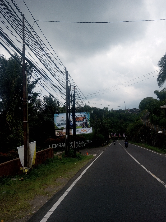 The image depicts an outdoor road scene under an overcast sky, not in Kalchini, India, but in an area of Indonesia, as indicated by visible Indonesian text on a billboard and sign ("LEMBAH PE MAI RESORT," "CIPANAS," Indonesian phone numbers).
The foreground shows a paved asphalt road with a solid white line marking the left edge and a dashed white line indicating a lane division further right. The road curves gently to the right as it recedes into the distance. On the left side of the road, multiple dark utility poles stand vertically, supporting a dense network of black electrical or communication cables strung overhead. Lush green vegetation, including tall, palm-like trees and dense foliage, borders both sides of the road.
A large billboard is prominently displayed on the left, behind the utility poles and vegetation. It features images of individuals riding all-terrain vehicles and displays text advertising "LEMBAH PE MAI RESORT" and "CIPANAS," along with phone numbers and a "90K" price indicator. Below this, a darker sign with white lettering partially reads "LEMBAH PE MAI RESORT CA CIPANAS." A white and yellow plastic sheet or debris is visible near the base of the leftmost utility pole. A small orange traffic cone is present on the grass verge near the billboard.
The right side of the road is bordered by a steep, heavily forested hillside. In the mid-distance on the road, three individuals are observed riding motorcycles away from the viewer; all are wearing helmets. Further up the distant hillside, a cellular tower or antenna structure is visible. The sky is uniformly grey with dense clouds.