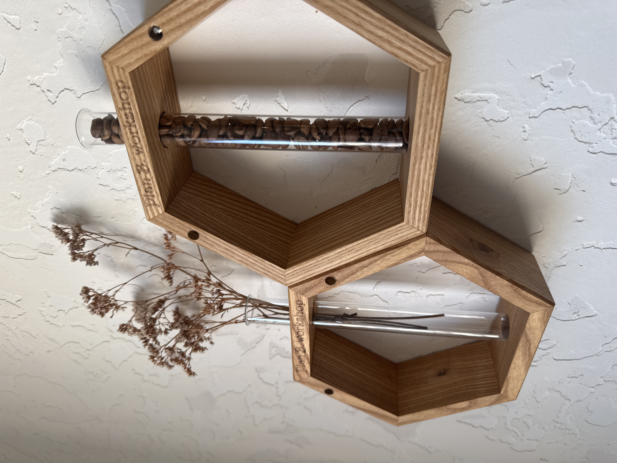 Two light brown hexagonal wooden frames are mounted on a textured white wall. The top frame contains a glass tube filled with coffee beans. The bottom frame contains a glass tube holding dried flowers. "Plan B. workshop" is etched on the side of each frame. The location is Chaikovsky, Russia.
