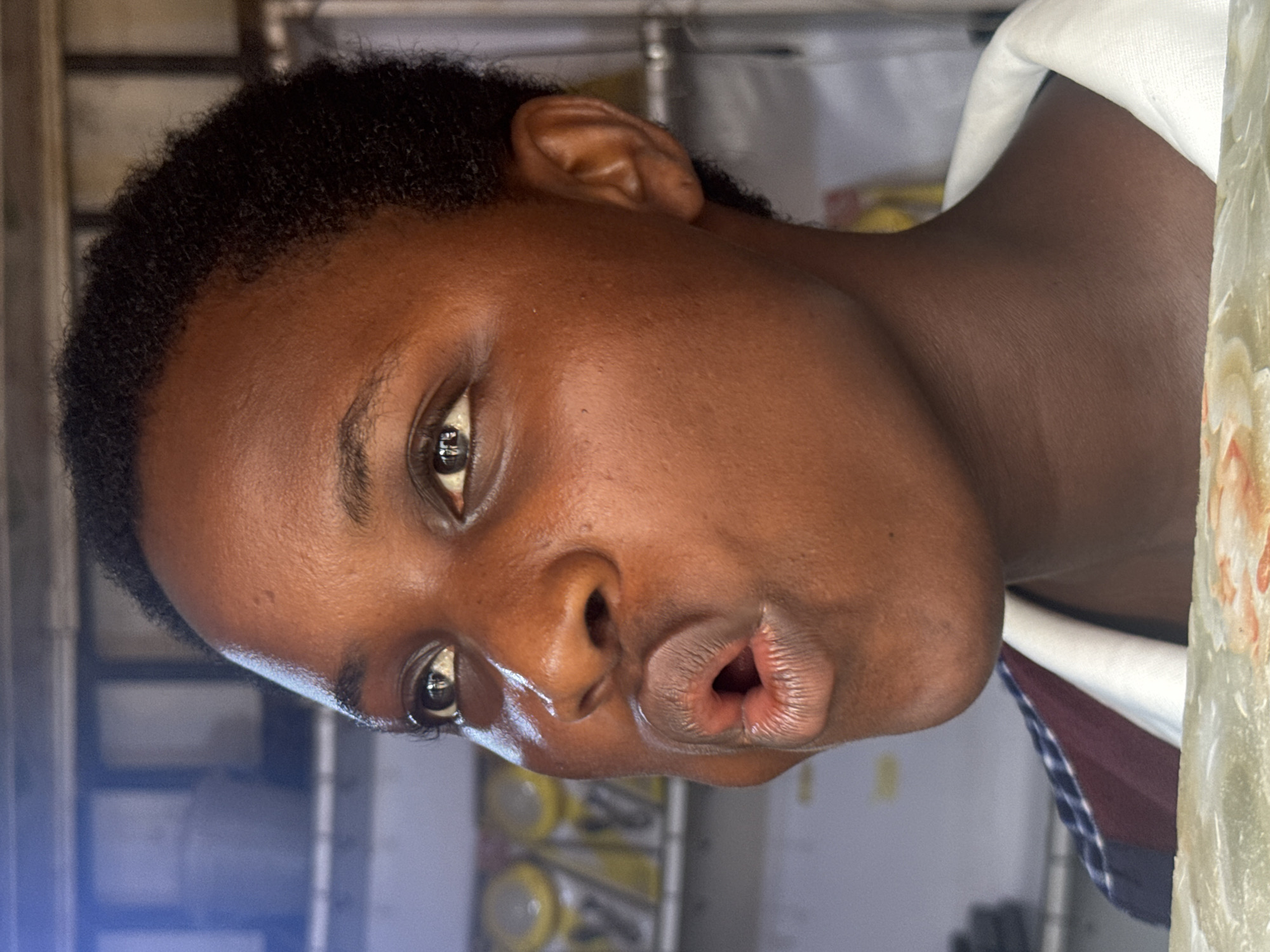 A close-up, eye-level, medium shot captures the head and shoulders of a young Black person in Ihiagwa, Nigeria. The individual has dark, short hair and medium-brown skin with a slightly dewy complexion. Their mouth is open in a puckered or surprised expression, with the lips pursed and slightly parted. Their eyes are open and looking upward and to the left, with dark irises and prominent eyelashes.
The background is out of focus, suggesting a shallow depth of field. On the left, a blurred shelf contains several yellow cylindrical objects, possibly bottles or containers. Further back, a dimly lit area shows shelving with what appear to be white rectangular objects and some bluish light. The person is wearing a white collared shirt with a dark, patterned neckline visible. The bottom of the image is cropped to show a textured surface, possibly a table or counter, with a faint floral pattern. The lighting creates highlights on the person's forehead, nose, and cheeks, indicating a light source from the front and slightly to the left.
