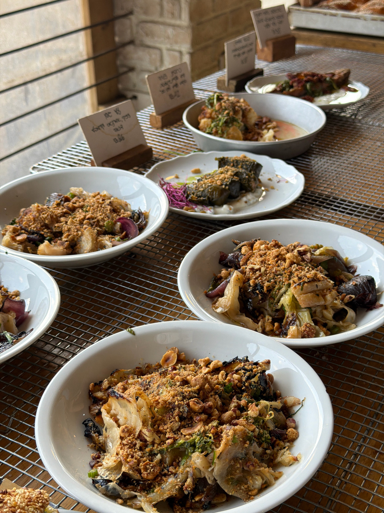 This image captures a food presentation, likely from a catering event or a restaurant with a buffet-style setup, at a location identified as Kfar Monash, Israel. The primary subjects are several white ceramic bowls filled with food, arranged on a metal cooling rack. The food appears to be a mix of roasted or grilled vegetables, possibly cabbage and eggplant, topped with a crumble of nuts and herbs. Small white signs with handwritten text are placed in front of some of the bowls, indicating menu items or pricing. The background shows a wooden slatted wall and a brick wall, suggesting an indoor or semi-outdoor dining area. The lighting is natural, coming from the left, casting soft shadows. There are no people visible in the frame.