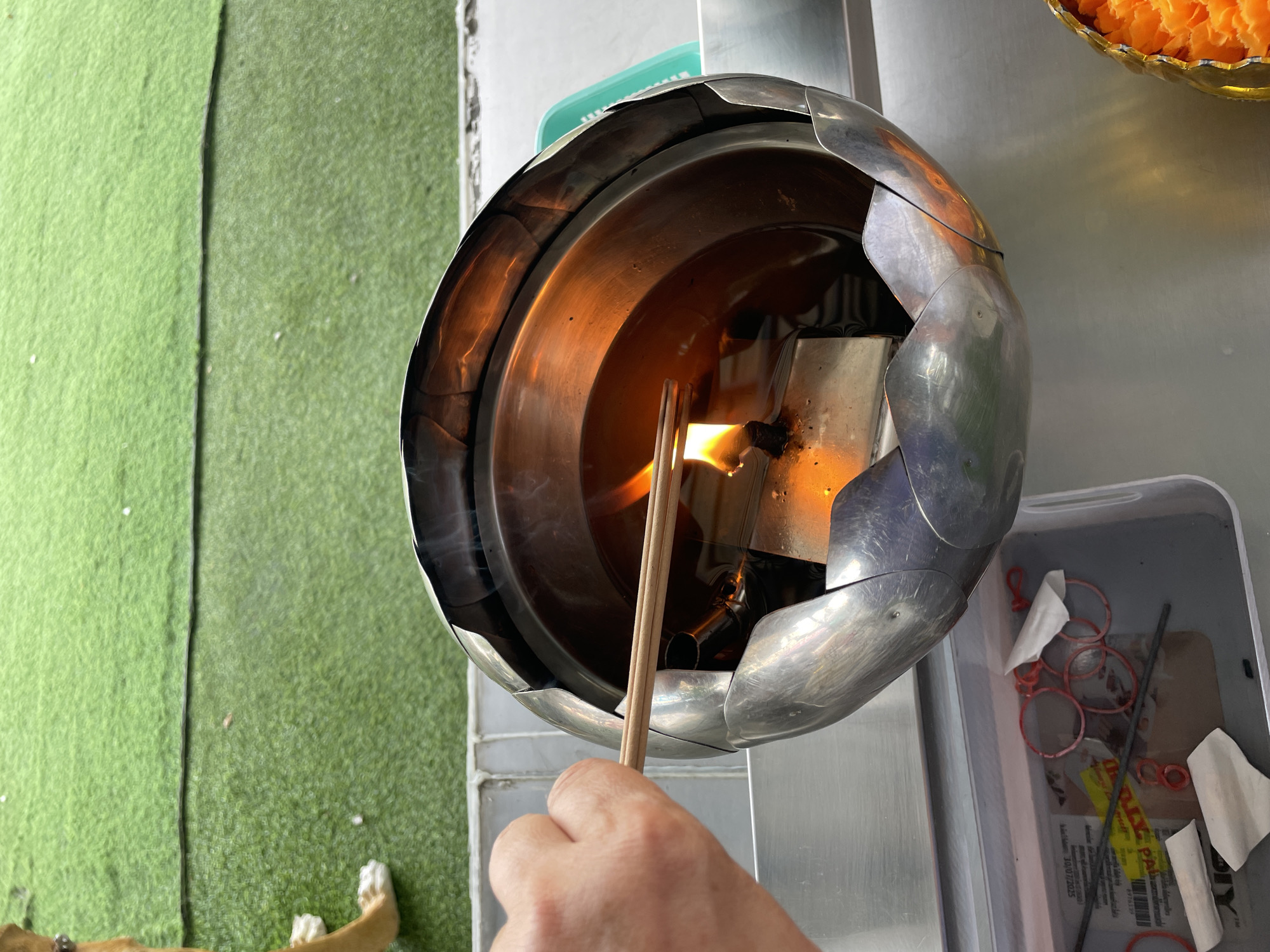An individual's hand holds two unlit incense sticks over a metal brazier filled with oil and a lit wick. The brazier is ornate, with petal-like protrusions around its rim. The background consists of green artificial turf, with a sliver of a dog's paw visible on the far left. The scene appears to be outdoors, possibly in a temple or shrine in Tambon Na Chom Thian, Thailand, given the presence of incense and the artificial turf often found in such locations. A grey metal surface runs horizontally across the bottom of the image, with a plastic container holding red rubber bands and a metal rod.