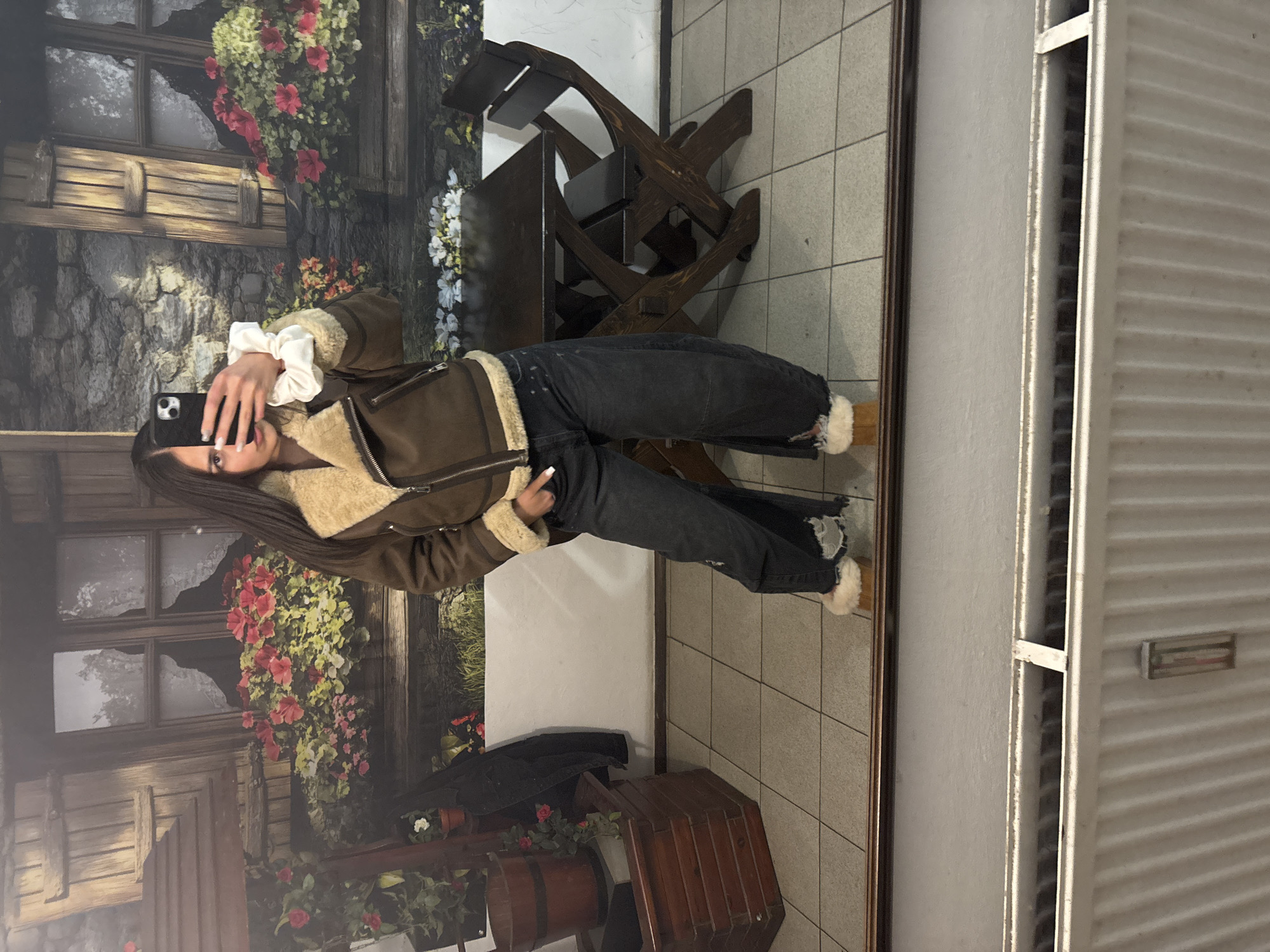 A woman takes a selfie in front of a wall mural depicting a building facade covered in flowers. She is wearing a brown jacket with fur trim and dark jeans. Behind her is a wooden table and chairs. To the right of the frame is a white door with horizontal slats. The location might be in Gabrovo, Bulgaria.