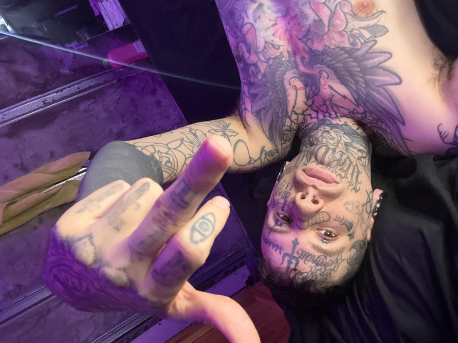 An individual, appearing male, is depicted in a high-angle, close-up photograph, lying on a dark surface, likely bedding, in an indoor setting. The ambient lighting casts a strong purple-magenta hue across the scene. The subject is extensively tattooed on the face, neck, chest, and arms. Facial tattoos include script and symbols, notably "MAMA" above the right eyebrow, a bandage design on the right cheek, and text resembling "GANG" on the left cheek. Multiple facial piercings are visible: several nostril studs, a septum ring, and a black ear gauge in the left ear. The subject's left hand is raised towards the camera, with the index and middle fingers extended and the ring and pinky fingers curled, revealing tattoos on the fingers including an eye symbol on the left middle finger. The individual is shirtless, displaying extensive chest tattoos, including a large winged creature design, and full sleeve tattoos on both arms. The subject maintains direct eye contact with the camera, with a neutral facial expression. The background is dimly lit and indistinct, with minimal identifiable objects: a folded light brown fabric, a pink plastic bottle, and a stack of items are visible on a dark surface to the right. There are no discernible elements within the image to specify a location within the city of Buenos Aires, Argentina.