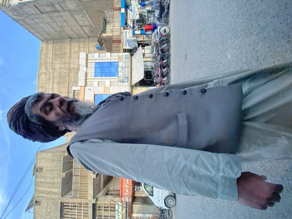 The image depicts an elderly man walking on a paved street in a commercial district of Kandahar, Afghanistan. The man, positioned off-center in the foreground, wears a dark, wrapped turban, a light-colored long-sleeved garment, and a dark grey button-up vest. He has a prominent grey beard, and his gaze is directed downward and to his left. The background features multi-story urban buildings under a clear blue sky. On the left, a white van is partially visible behind the man, parked next to a building with a grey concrete facade and visible balconies. On the right side of the street, a dense arrangement of motorcycles and at least one bicycle is parked along the sidewalk, in front of various storefronts. One building in the background displays reflective blue-tinted windows, while another features a white awning. The street surface appears worn and is visible in the lower portion of the frame. No direct interactions between individuals are discernible.