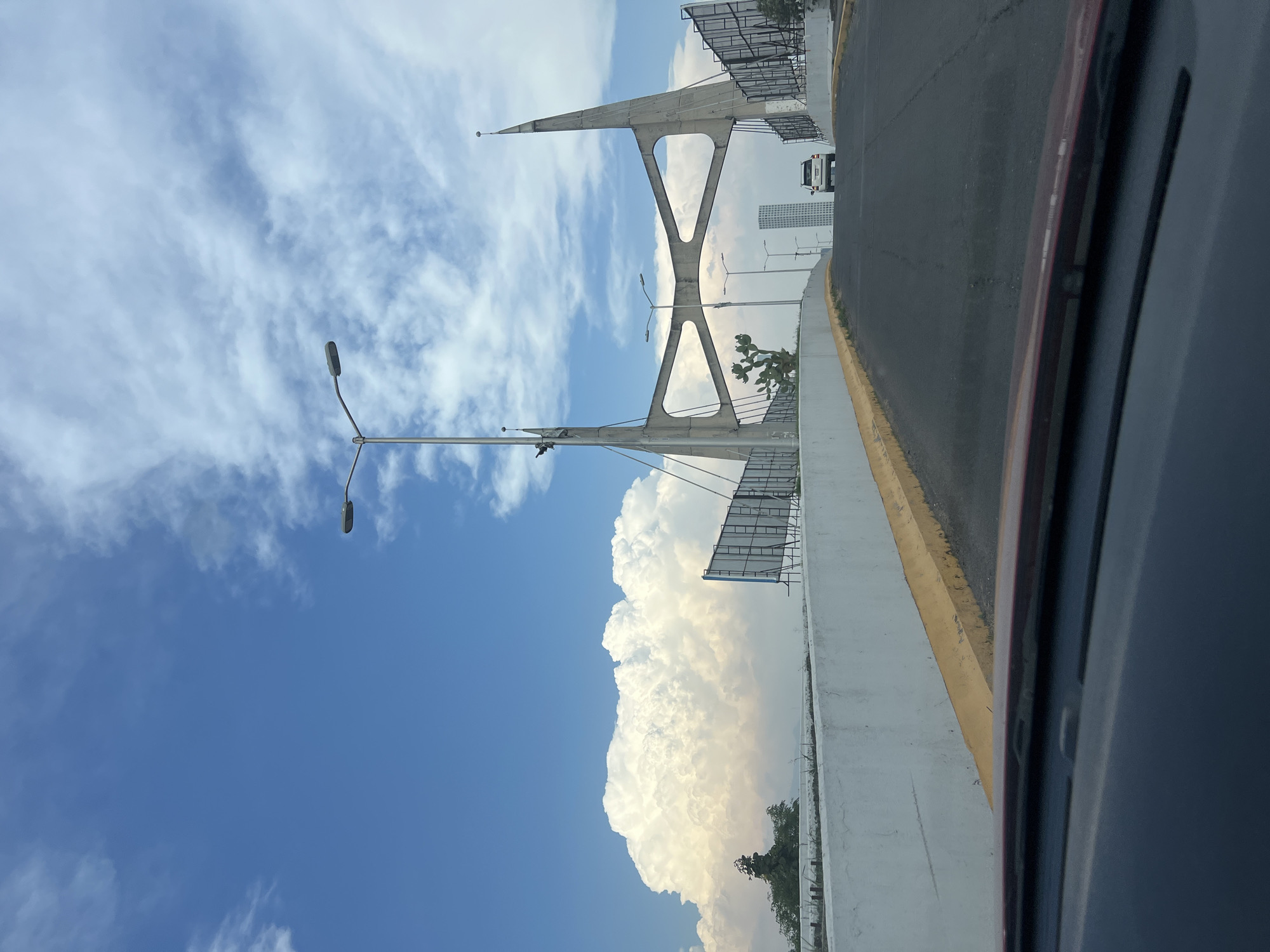 A view from inside a moving vehicle, likely on a bridge, looking across a road towards a large, billowing cloud formation under a blue, partly cloudy sky. The architecture of the bridge includes a concrete structure with an inverted triangle design. There are lampposts along the road, and billboards are visible attached to the bridge structure. In the distance, a tall building and a vehicle can be seen. The scene appears to be near San Bernardino Tlaxcalancingo, Mexico, based on the architectural style and environment. The road has a yellow curb. The vehicle's interior is visible along the right edge of the frame.