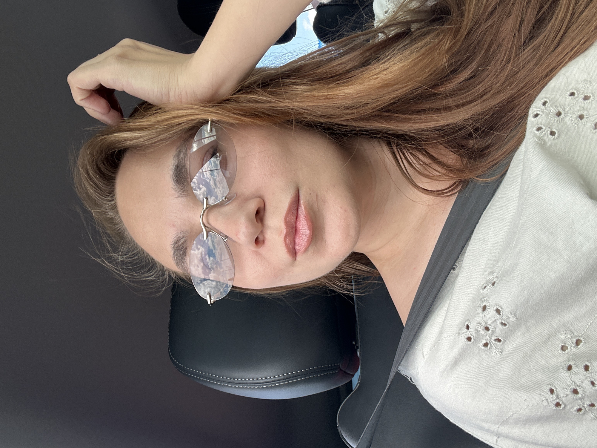 The image shows a woman in the passenger seat of a car, with her hand resting on her head. She is wearing diamond-shaped glasses that reflect the sky. The background is dark, suggesting the interior of the car, with a hint of a window and sky visible in the upper right. The woman has long, reddish-brown hair and is wearing a white, embroidered top. The car appears to be on a road, as suggested by the reflection in her glasses and the implied movement. The location is Ferma Sovkhoz Balykleyskiy, Russia.