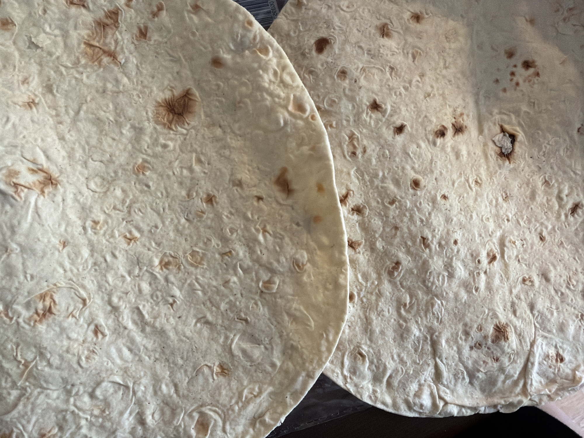 The image shows two flatbreads, possibly lavash or a similar type of thin, unleavened bread. They are light in color with small brown spots, likely from cooking. The bread on the left is closer to the camera, partially overlapping the one on the right. The background is a dark, flat surface. There is no information in the image that relates to the city of Saint Petersburg, Russia.