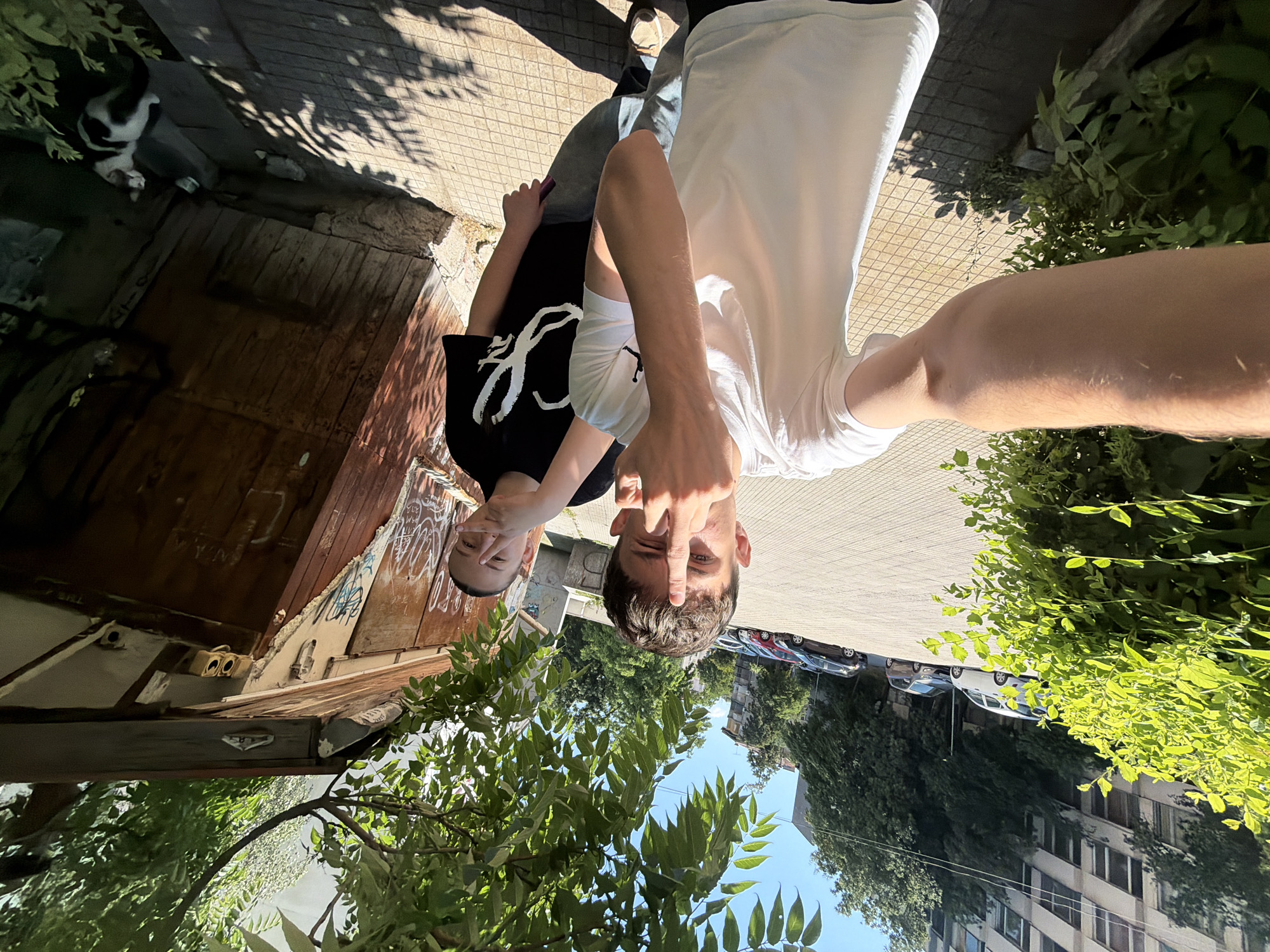 Two young individuals are captured in a selfie-style photograph on a sunlit day in Plovdiv, Bulgaria. The foreground is dominated by a person wearing a white t-shirt and jeans, extending their arm towards the camera and displaying a middle finger. To their right stands another individual in a black t-shirt with a white graphic, also making a gesture with their hand. They are positioned on a paved walkway with a distinct geometric pattern, bordered by lush green foliage on the left. In the background, several parked cars are visible on a street adjacent to a multi-story residential building. To the right of the individuals, a rustic, weathered wooden structure with graffiti adorns its surface, and a black and white cat is present near its base. The scene suggests an outdoor urban environment, possibly a residential area or a public space.