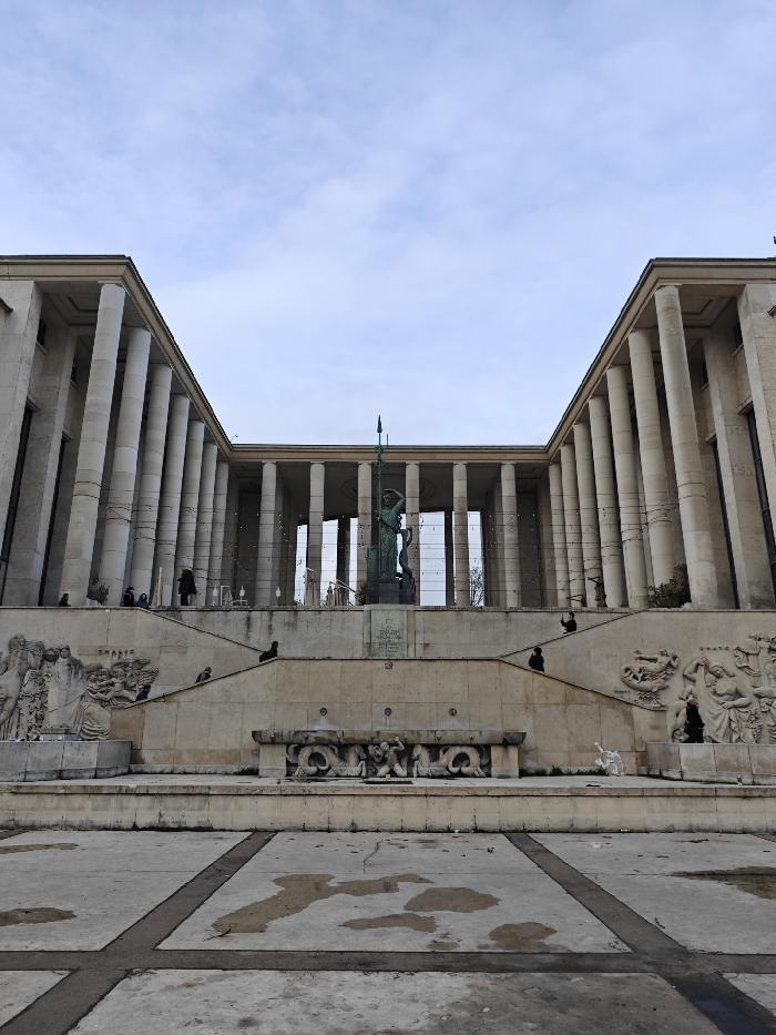 The image displays a wide, frontal view of the Palais de Chaillot in Paris, France, specifically the section facing the Trocadéro Gardens and Esplanade. The monumental structure consists of two symmetrical, curved wings featuring extensive colonnades of numerous tall, light-colored columns supporting a flat roofline. A recessed central section between the wings also exhibits columns and large openings, strung with multiple horizontal lines of small, white decorative lights.
Centered in the foreground of the building is a large, dark-colored bronze statue, possibly titled "La France renaissante," depicting a standing figure, potentially female, holding a long spear or staff vertically in its left hand. This statue rests on a multi-tiered stone pedestal with an inscription.
Below the main building and statue, a series of wide, light-colored stone terraces descend towards the viewer. Two broad, symmetrical staircases diverge from the lower foreground, ascending to meet near the central statue. The walls flanking these staircases and terraces are adorned with large, intricate bas-relief sculptures depicting various human figures and allegorical scenes. Directly in front of the central statue at a lower level, a decorative stone structure resembling a dry fountain basin is visible, featuring carved marine or mythical creatures.
Several individuals are sparsely distributed across the scene. Dark-clad figures are visible standing and sitting on the upper terrace near the building's columns and on the mid-level staircases.
The immediate foreground consists of a flat, light-grey paved area, divided into sections by darker lines. Numerous irregular, dark, wet-appearing patches or stains are present on this paving. The sky above is overcast, providing soft, diffused lighting and no distinct shadows.