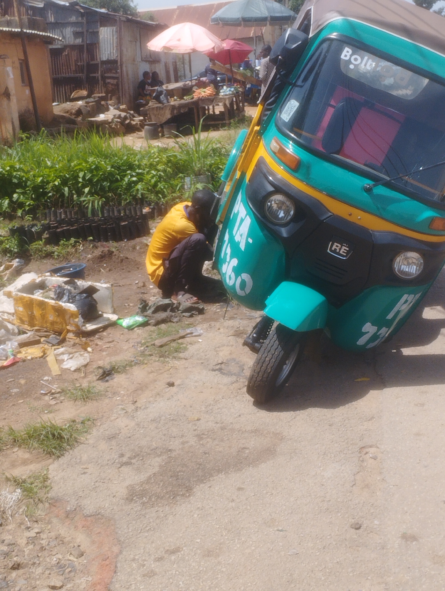 The image depicts an outdoor scene, likely in Jos, Nigeria, given the context. In the foreground, a teal and yellow auto-rickshaw, also known as a "Keke," is prominently displayed, tilted slightly to the right. White lettering on the vehicle reads "Boltu," and other illegible text is present on its side. A person wearing a bright yellow t-shirt and dark pants is crouched next to the Keke, appearing to be working on or attending to it. Scattered debris and trash are visible on the ground around the person and the vehicle, including a discarded yellow and white box, black garbage bags, and some green packaging.
In the background, several market stalls with corrugated metal roofs and varying building materials suggest a bustling market or a semi-permanent settlement. People are seen in the background, some seated at the stalls, with items like produce or goods displayed. Umbrellas, one pink and one green, are also visible, indicating it might be a sunny day with vendors seeking shade. The ground is a mix of dirt and rough paving, typical of an outdoor market or street in this region. The overall impression is one of everyday activity in a busy, possibly informal, commercial area. The lighting suggests it is daytime.