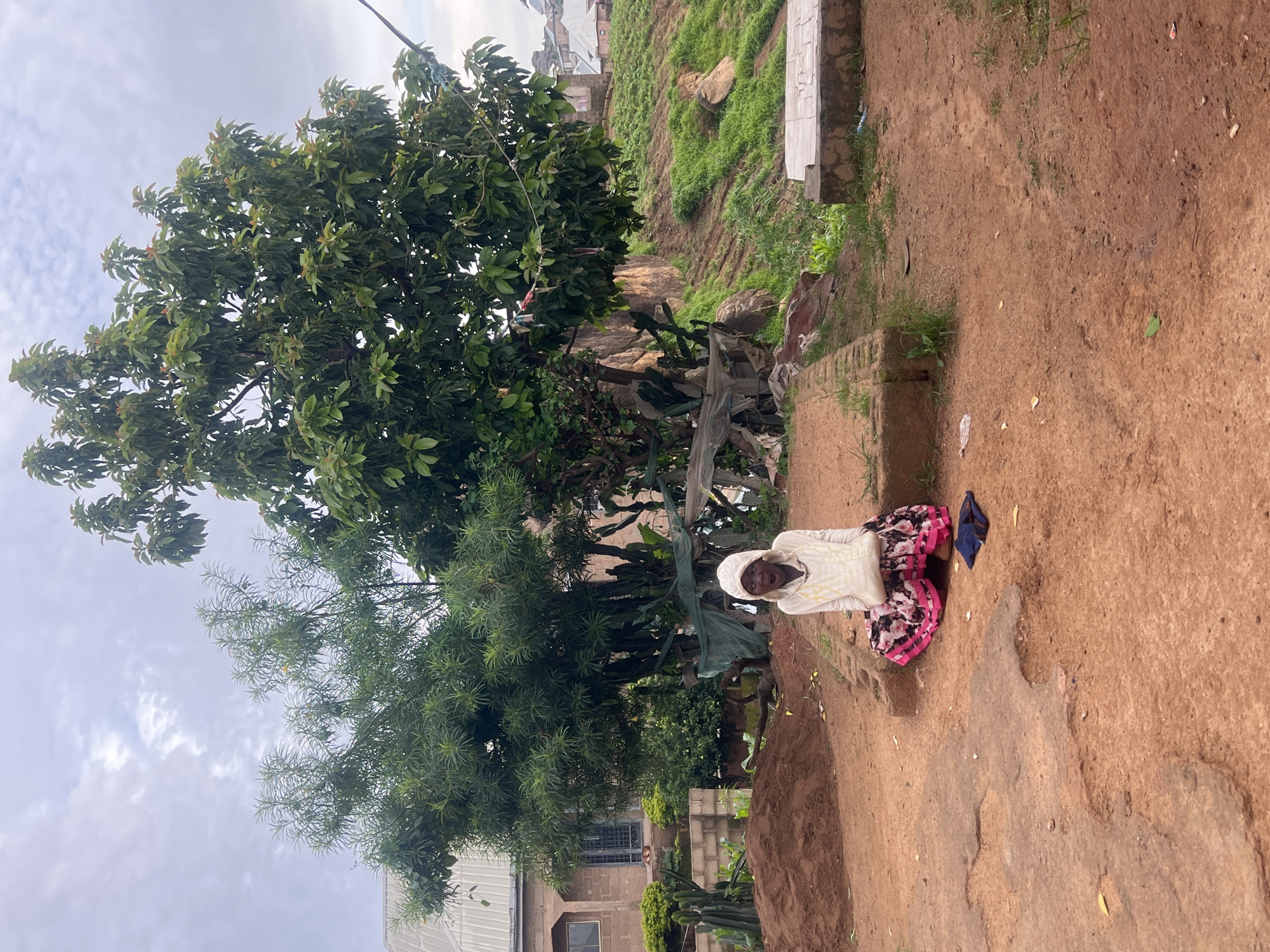 A person sits on a raised concrete platform in a dirt clearing. The person is wearing a white hooded garment and a long floral skirt. A dark item of clothing is on the ground to the person's right. Behind the person is a large green tree and a hill covered in grass and rocks. To the left of the tree is a brick building with windows. The ground is a reddish-brown dirt. The sky is overcast and cloudy. The location is a residential area in Jos, Nigeria.