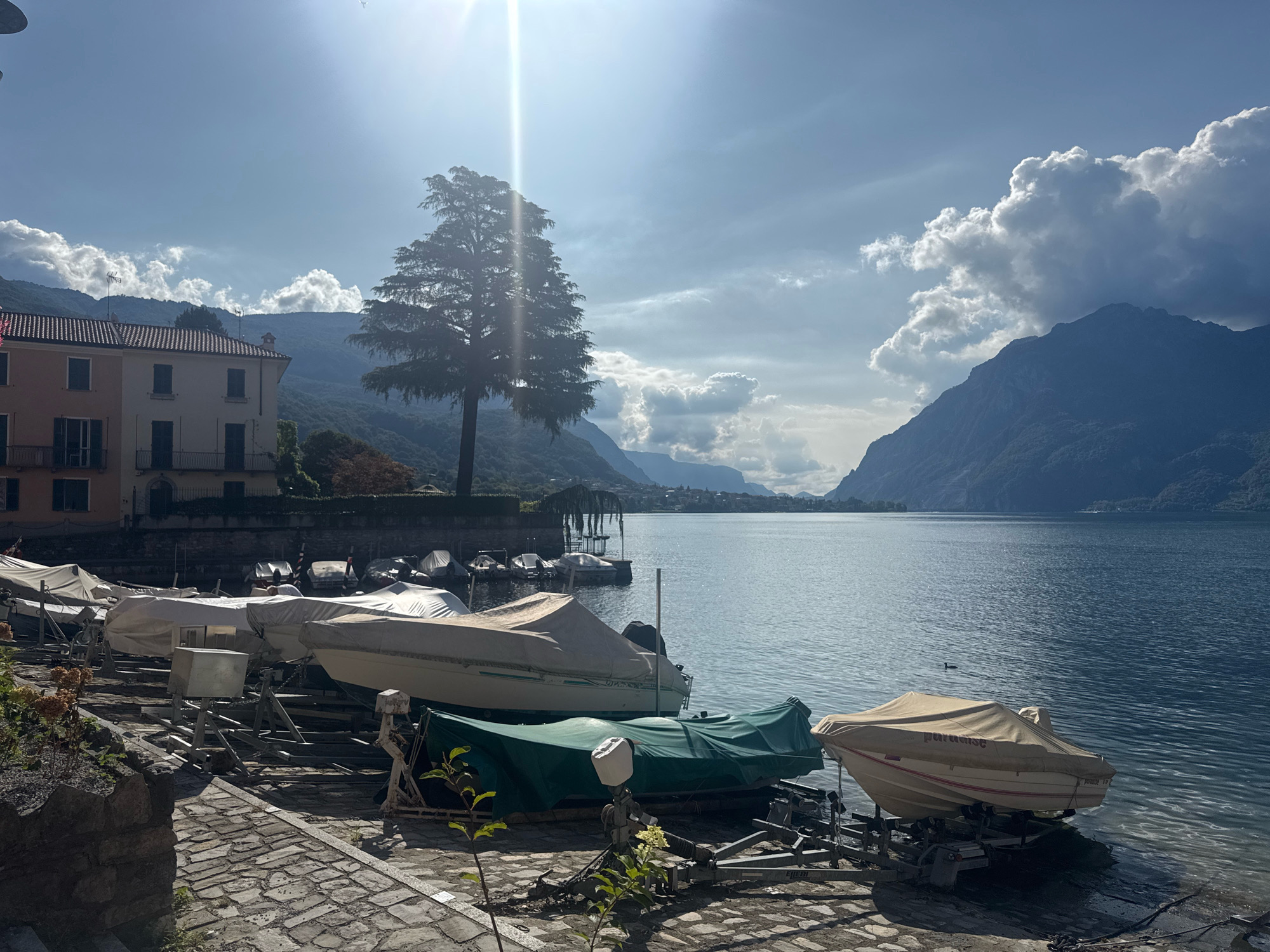 The image shows a waterside view in Mandello del Lario, Italy, with several boats covered and parked on trailers along the shore. A large tree stands in the foreground, silhouetted against a bright sky with sun flares. In the background, a body of water stretches out towards mountains under a partly cloudy sky. A building with a light-colored facade and dark windows is visible on the left. The ground is paved with cobblestones.