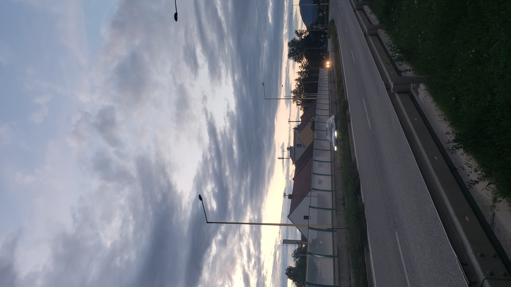 The image captures a twilight scene on a road in Prešov, Slovakia. A highway with guardrails runs diagonally through the frame, with a car in motion on the right lane, its headlights illuminated. To the left of the highway, a sound barrier is visible, separating the road from a row of houses with red-tiled roofs and several antennas. A tall streetlamp with a yellow pole stands prominently in the foreground. The sky is filled with dramatic, dark grey clouds, interspersed with lighter patches of sky indicating the fading light of dusk. Trees are visible in the distance, some silhouetted against the sky. The overall atmosphere is one of approaching evening in a suburban or semi-urban environment.