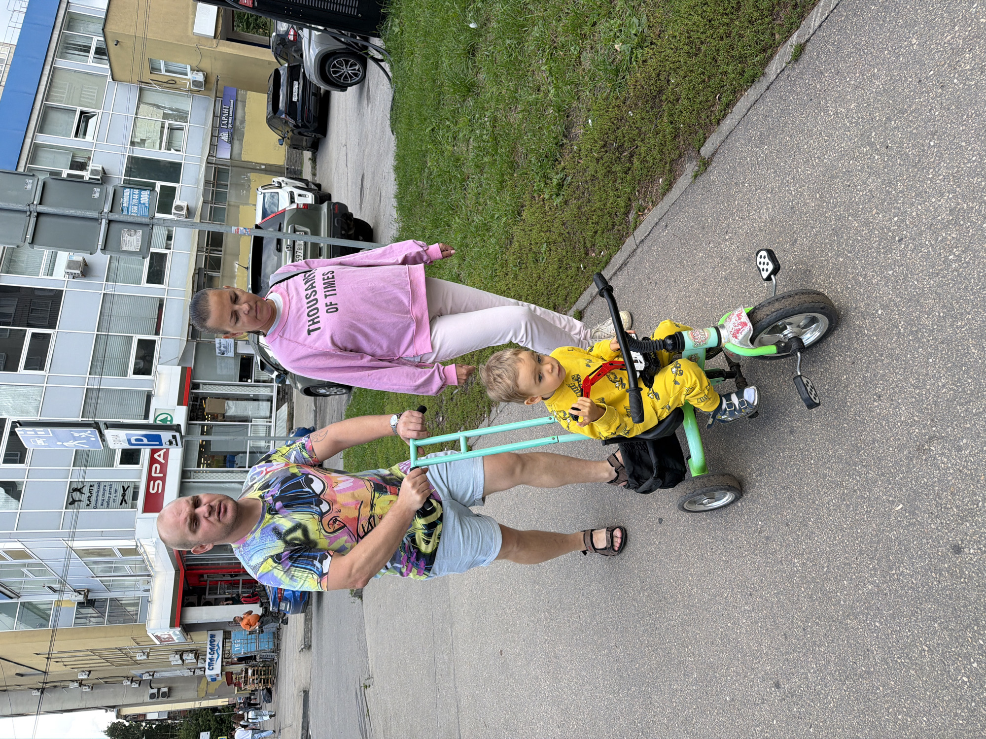 The image depicts a street scene in Nizhny Novgorod, Russia, on a cloudy day. A bald man, wearing a colorful graphic t-shirt and light blue shorts, pushes a teal and green children's tricycle with a young boy in a yellow jumpsuit. Beside them, a woman in a pink sweatshirt and light pink pants walks alongside. The tricycle has a black storage bag attached. In the background, a modern building with numerous windows dominates the scene, with signs for "SPA" and "KARATE" visible. Several parked cars are also present on the street, and patches of green grass border the sidewalk. The overall atmosphere suggests a casual outing in an urban environment.