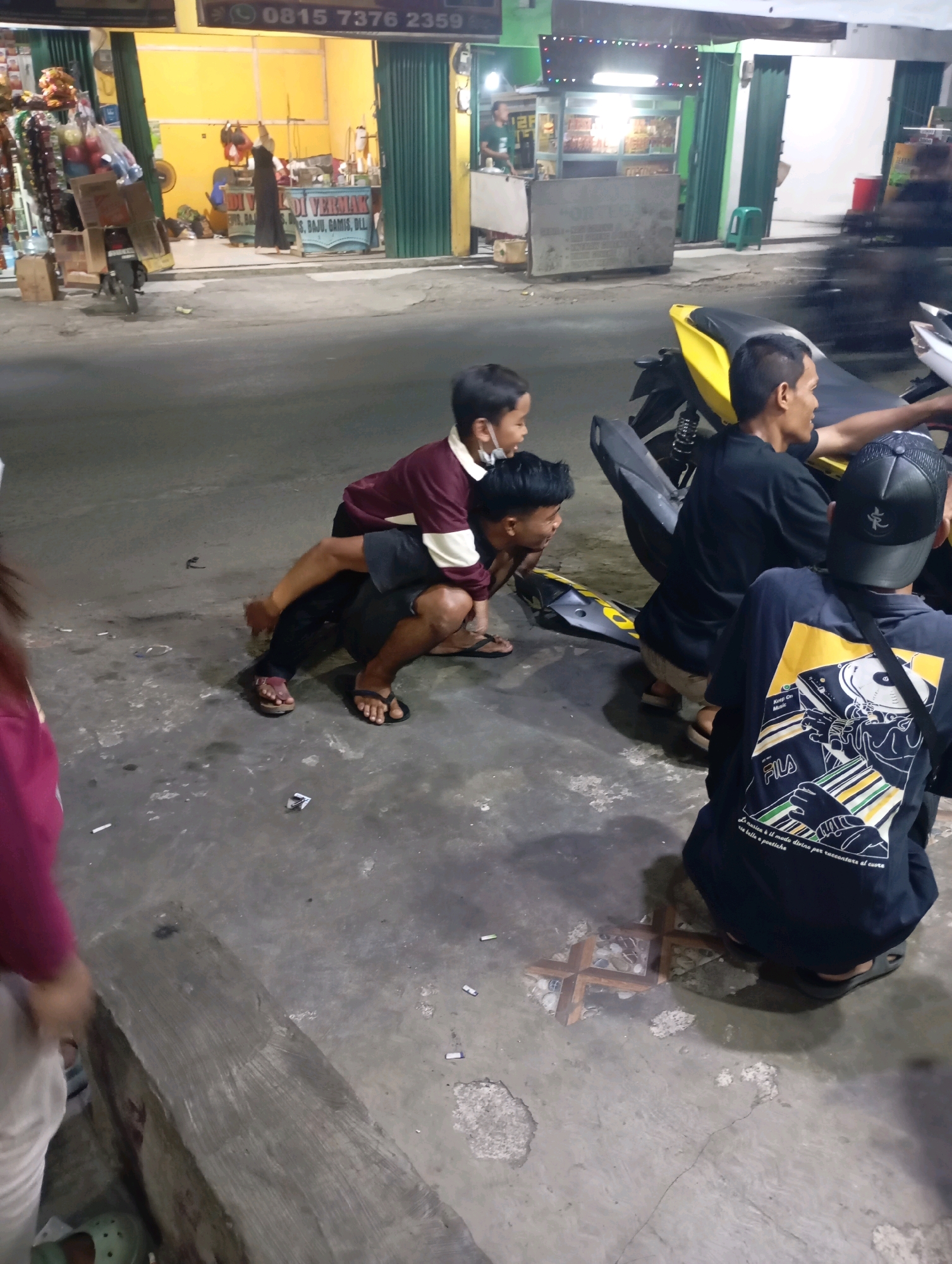 The image depicts a group of individuals in what appears to be an outdoor urban setting at night, likely in Babelan, Indonesia. In the foreground, a person wearing a dark t-shirt with a graphic design and a Fila logo is crouched down. To their left, another person in a black t-shirt is seated on a yellow motorcycle. A young child, wearing a maroon and white shirt, is being held from behind by an adult in a dark shirt and shorts. This adult is also crouched down. The environment is paved with concrete, and scattered on the ground are what appear to be cigarette butts and small pieces of debris. In the background, there are storefronts with green roll-up doors and illuminated interiors, suggesting a commercial area. One storefront has text that reads "ADI VERMAK" and mentions "S. BAJU, GAMIS, DLL." Another storefront displays food items. The overall scene suggests a casual gathering or stop in a public area.