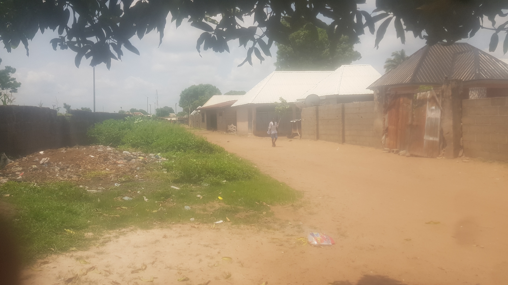 The image depicts a dusty, unpaved street in what appears to be a rural or semi-urban setting. The ground is a light brown dirt, with visible tire tracks and scattered debris. To the left, there is a large pile of garbage and vegetation, suggesting a lack of proper waste disposal.
Several buildings line the street. They have simple construction, with brick walls and corrugated metal roofs, some painted white. One building in the mid-ground has a person walking in front of it. This individual appears to be a young male wearing a light-colored shirt and dark shorts, walking away from the camera. Other structures visible include a fenced yard with a large, rustic metal gate and a building with a conical, metal roof. The overall architecture suggests a warm climate.
The sky is overcast with clouds, indicating it might be a cloudy day or potentially the early morning or late afternoon. Lush green foliage, including trees and bushes, is visible in the background and framing the top of the image, suggesting a somewhat tropical or sub-tropical environment. The scene conveys a sense of everyday life in a community that may be underserved by infrastructure. There is no visible text in the image.