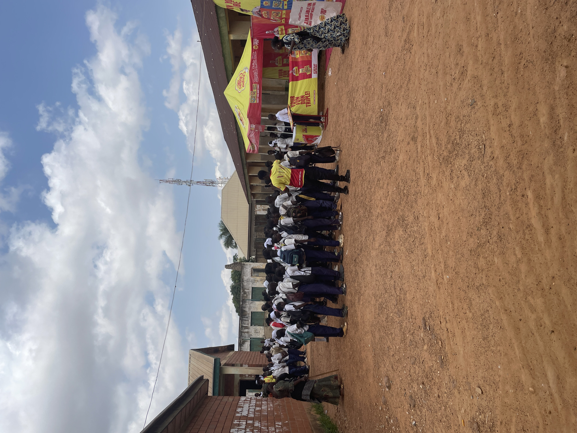 This image depicts a sunny daytime scene with a group of schoolchildren lined up outdoors. The children, dressed in uniforms, appear to be attending an event under a large tent branded with "Chupa Chups." The tent features colorful banners advertising the candy brand with slogans like "Stretch. Twist. Toss" and "YOUR FUN DON TRIPLE!" Text on the banners also mentions "AVAILABLE IN ANY SHOP NEAR YOU."
Behind the tent, there are buildings, possibly a school, with a red brick structure visible on the left. In the background, a cell tower stands against a bright blue sky dotted with white clouds. The ground is made of reddish-brown dirt. A woman in a patterned dress stands near the tent, interacting with something or someone out of frame. The overall atmosphere suggests a promotional event or school activity. The lighting and clear sky indicate it's a fair weather day, likely mid-morning or afternoon. The presence of the Chupa Chups branding and the uniformed children suggests a brand activation event aimed at students.