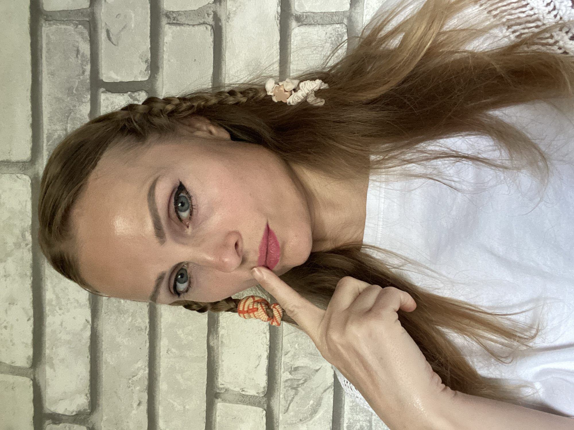 A medium close-up frontal shot shows a white woman with blonde hair styled in two braids with decorative ties. She is wearing a white shirt with lace trim and has makeup on her eyes and lips. Her right index finger is touching her upper lip, and she is looking directly at the camera with a neutral expression. The background is a light-colored brick wall. The image does not contain any identifiable landmarks or information that would place it in the city of Міханавіцкі сельскі Савет, Belarus.