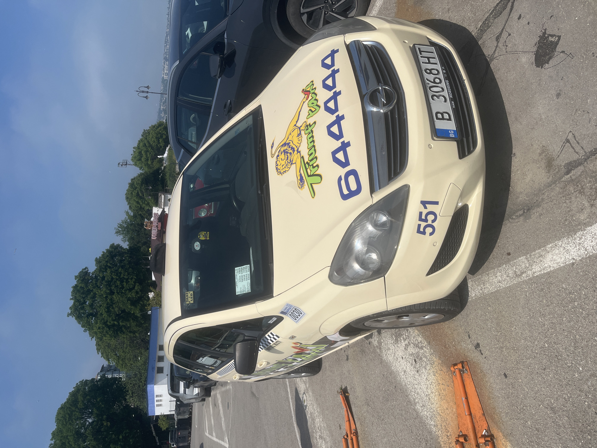 A beige Opel car, identifiable as a taxi, is prominently featured, parked outdoors on an asphalt surface. The car is angled, with its front facing towards the right. On the hood, a vibrant graphic displays a yellow lion alongside the green and yellow text "Trumf Taxi" and the large blue digits "644444", likely a phone number. The taxi's identifier number "551" is also visible in blue on the front bumper, below the grille.
The Bulgarian license plate "BG B 3068 HT" is clearly mounted on the front of the vehicle, with "TEAM NORDRING" printed below it. Other visible text includes "0030" on a sticker near the front fender, "TAXI LED" on a small sign, and a "SMILES" sticker on the upper corner of the windshield.
Behind the taxi, a dark-colored car is partially visible. The ground is asphalt with white parking lines and some dark stains, while an orange hydraulic jack or similar tool rests near the front wheel. In the background, green trees and distant buildings are visible under a bright, clear blue sky, indicating daytime. The scene depicts a parking area, consistent with the provided location context of Varna, Bulgaria.