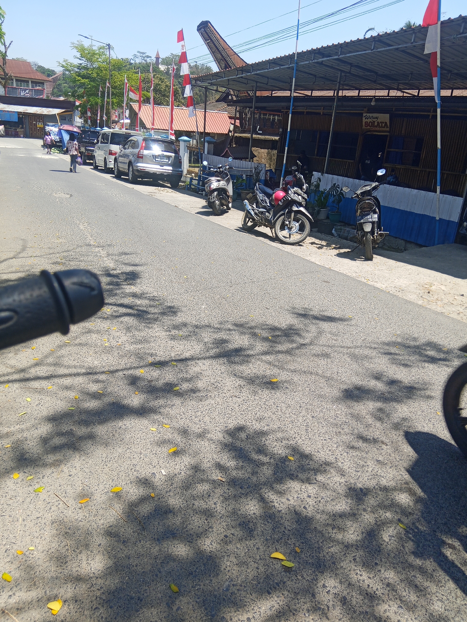 In Makale, Indonesia, a street scene unfolds on a sunny day under a clear blue sky. The road is paved, with fallen yellow leaves scattered across its surface. Shadows of trees are cast onto the asphalt, creating a dappled effect. On the right side of the road, a building with a corrugated metal roof, supported by bamboo poles, houses a restaurant labeled "WARUNG SOLATA." A blue and white striped facade runs along the front of the building. Several motorcycles are parked in front of the restaurant, including a silver and red motorcycle and a black motorcycle. A white van and a silver SUV are parked further down the street on the left. Two individuals are walking on the sidewalk on the left side of the road, one carrying a blue umbrella. Red and white Indonesian flags are displayed on poles along the street. The foreground of the image is dominated by a blurred black object, possibly a handlebar.