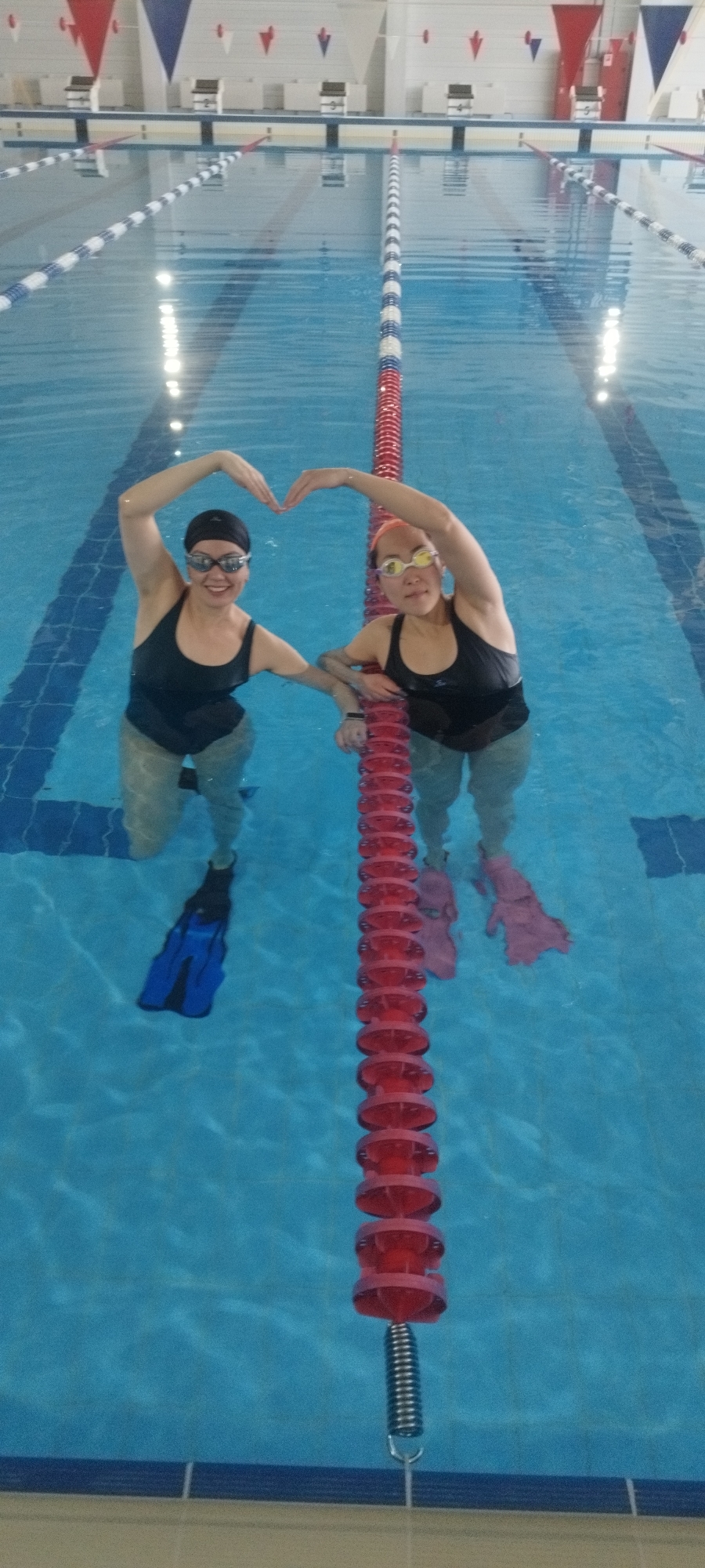 Two women are standing in a swimming pool, making a heart shape with their arms above their heads. The woman on the left is wearing a black swim cap, goggles, a black swimsuit, and blue fins. The woman on the right is wearing goggles, a black swimsuit, and pink fins. They are both standing near a lane marker in the pool. The pool has multiple lanes, indicated by lane markers, and there are starting blocks visible in the background. Red, white, and blue pennants hang above the starting blocks. The pool is located indoors. The location is a swimming pool but there is no location found with the city of Yakutsk, Russia.