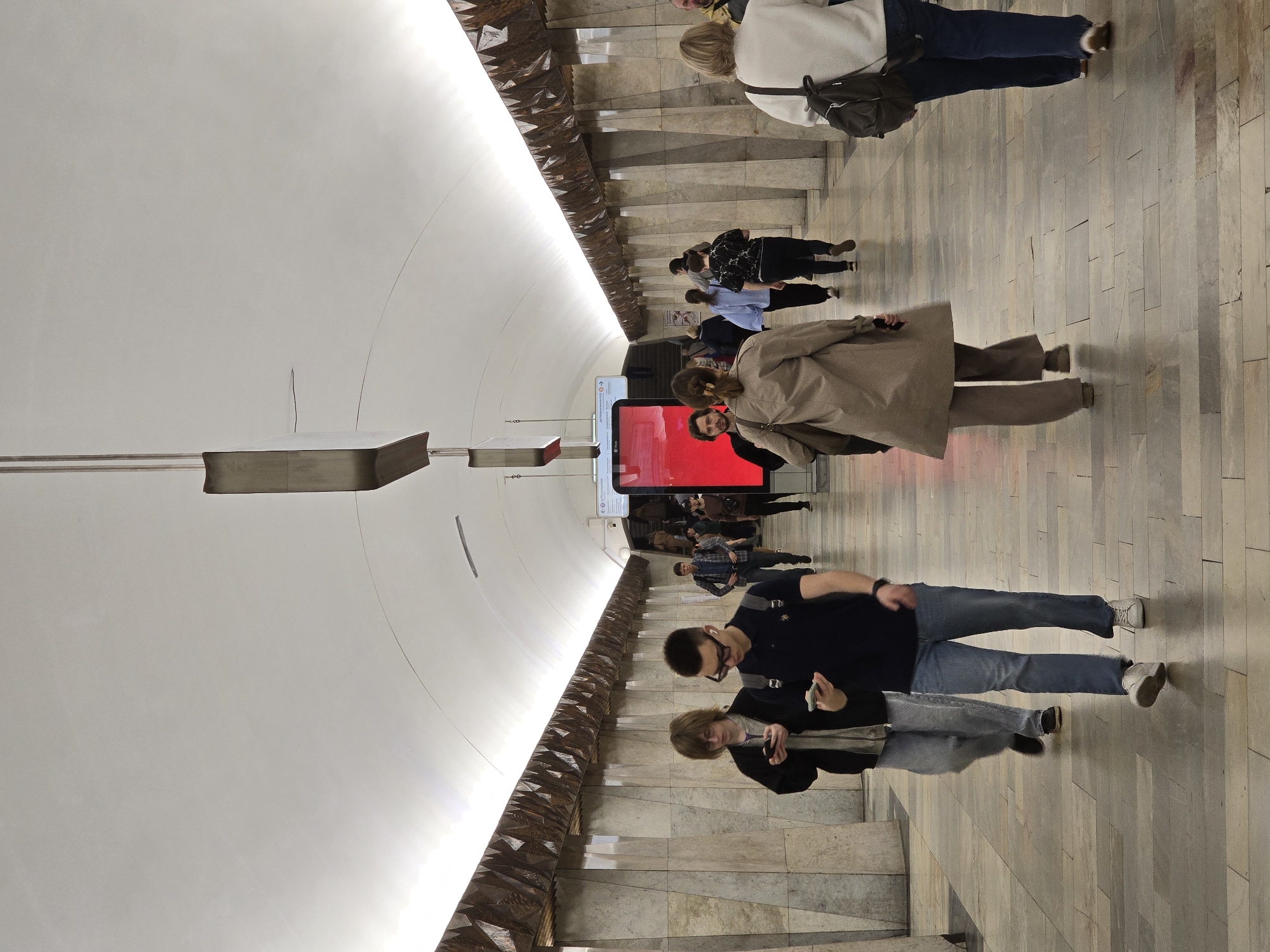 Here is a description of the image:
The image captures an underground public space, potentially a metro station in an unknown city, featuring a group of people navigating a descending staircase. The architectural design includes a distinctive, rounded ceiling that curves downwards and is adorned with geometric, decorative elements along the edges where it meets the wall.
Several individuals are moving towards the bottom of the staircase, each engaged in their own activity: a man wearing a black shirt and jeans is looking at his phone; a woman wearing a tan trench coat is walking downwards with her hand in her pocket. The lighting is bright, accentuating the ceiling's design and highlighting the movement of people in the public space. The scene is crowded with commuters, suggesting a busy transportation hub. A tall digital advertisement displays a large red screen to the bottom of the stairs.