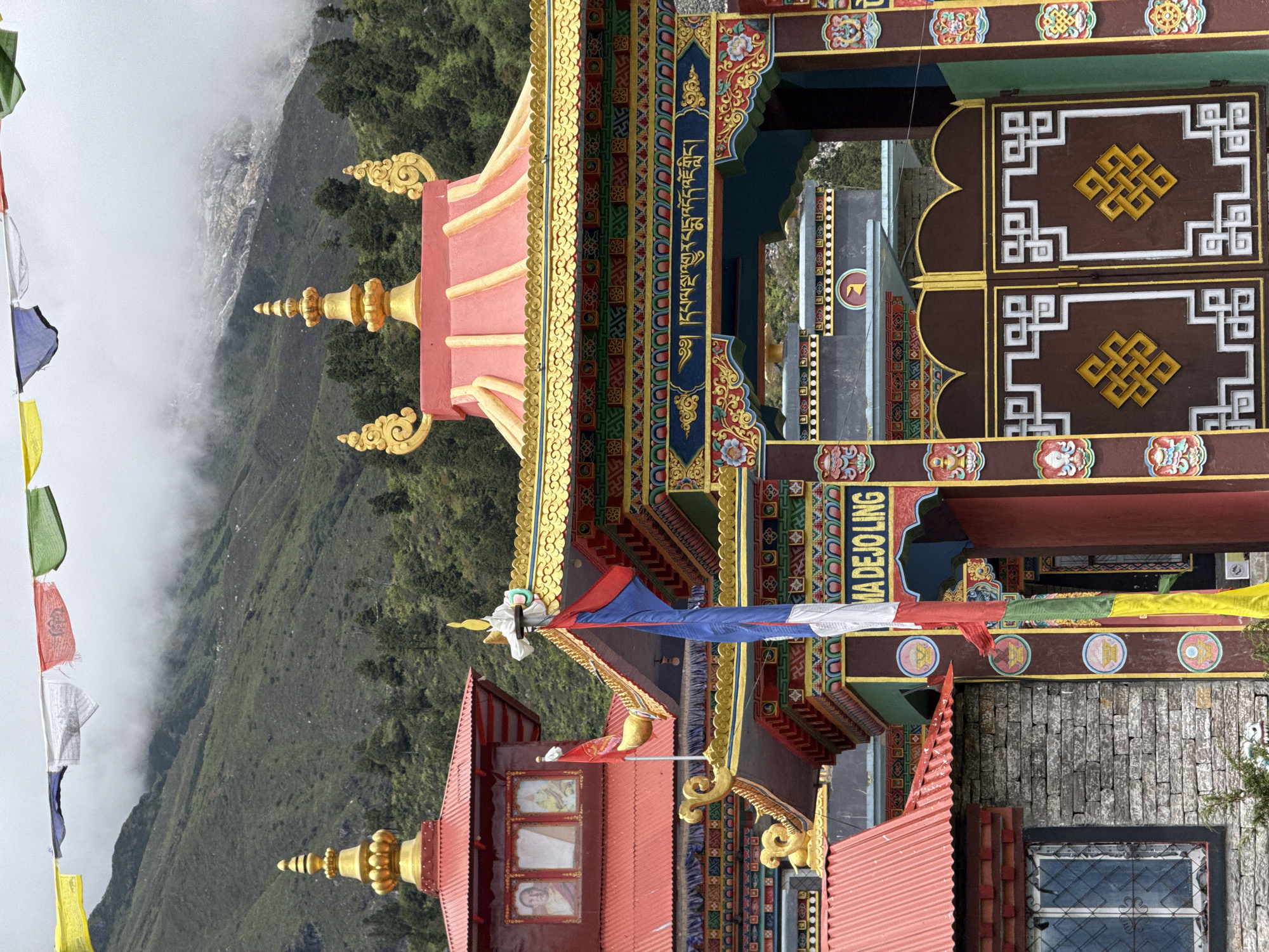 This is a medium shot, eye-level photograph of the entrance to the Gekay Ling Monastery in Samagaon, Nepal. The monastery's architecture is predominantly red and gold, with intricate decorative details. Prayer flags in various colors are strung across the top of the frame, with a lush, green mountainside in the background, partially obscured by mist. The entrance gate is elaborate, featuring a golden symbol on a dark brown background. To the left of the gate, a stone building with a red roof and a window with a metal grate is visible. The overall scene conveys a sense of spiritual and cultural significance, set against a dramatic natural landscape.