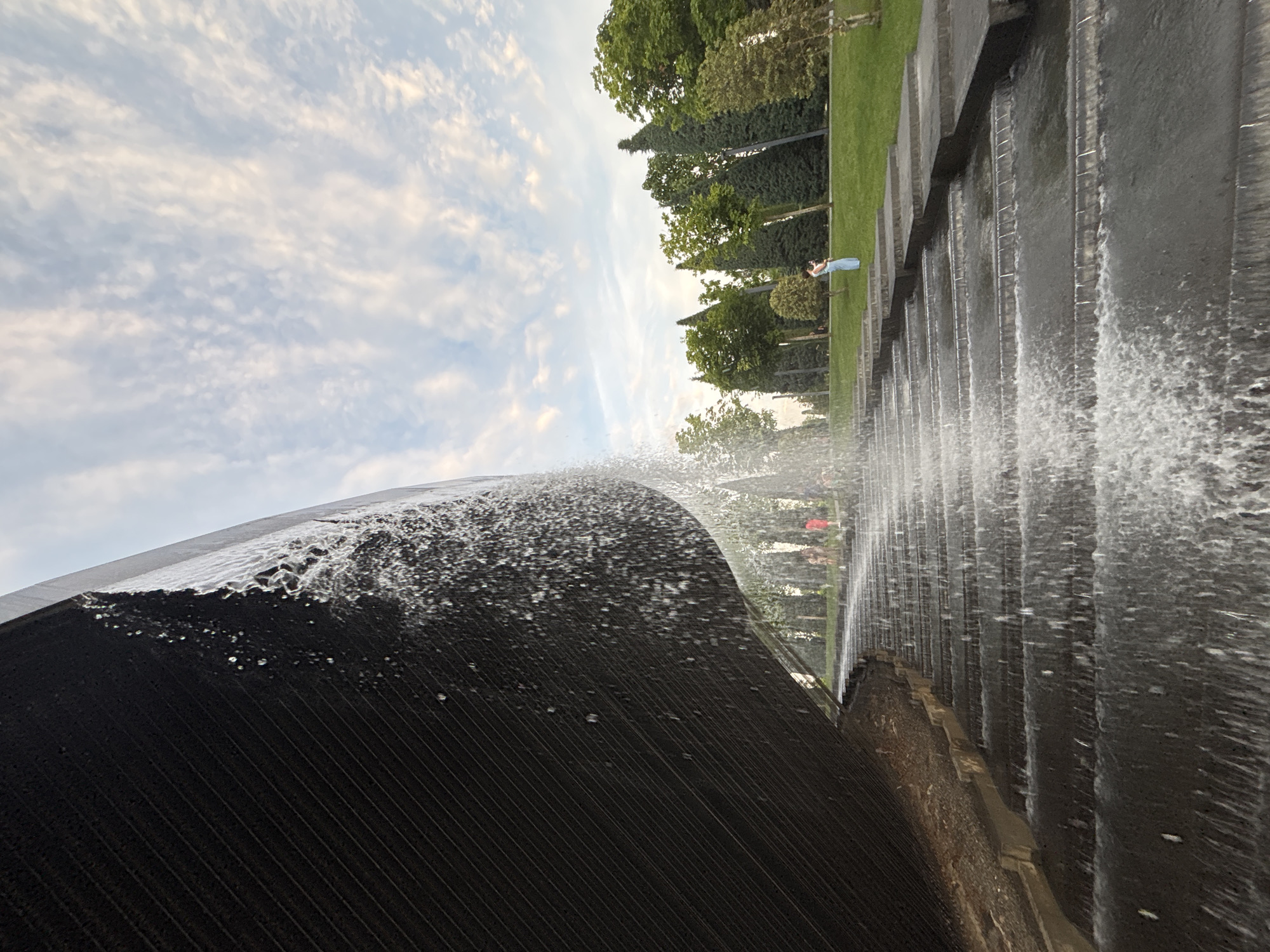 The image depicts a stepped water feature in a park setting. A large, dark, textured surface, likely a wall or structure, is visible on the left, with water cascading down in front of it. The water flows over a series of concrete steps, creating a textured effect with foam and spray. In the background, a lush green lawn is dotted with various trees, including tall, slender evergreens and broader deciduous trees. The sky above is a soft blue, with scattered, wispy white clouds.
A few people are present in the mid-ground, standing on the grass and on some of the upper steps of the water feature. One individual, wearing a light blue outfit, is standing on the grass in the middle right of the frame, looking towards the right. Several other figures, appearing as casual park visitors, are visible on the steps further back and to the left. The overall scene suggests a tranquil park environment, possibly in the late afternoon or early evening, given the quality of the light. The location is identified as the Public Garden "Solnechny Ostrov" in Krasnodar, Russia.