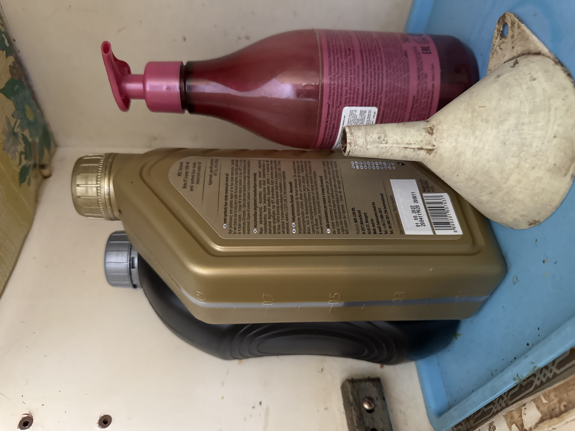 The image displays a close-up, eye-level view of several items stored on a light blue plastic tray, which itself is resting on a surface that appears to be a shelf or cabinet. The background is a light-colored, textured surface, possibly painted wood or plastic, with some faint floral pattern visible at the top.
Prominently featured are three containers. On the left is a black plastic bottle with a silver cap, mostly obscured from view. To its right is a gold-colored plastic bottle with a gold cap. This bottle has a label with text in multiple languages, including English, Hungarian, and German, indicating it contains "MOL Dynamic Moto 4T Scooter 10W-40 semi-synthetic four-stroke motorcycle motor oil." A clear blue stripe runs vertically along the side of the bottle, with visible fill lines marked at 0.7, 0.5, and 0.3. To the right of the oil bottle is a pink, translucent plastic bottle with a pink pump dispenser. The label on this bottle is mostly illegible due to blur and text size, but some French text is visible, along with the words "FORMULE PROFESSIONNELLE."
In the foreground, partially obscuring the bottom of the oil bottle and the pink dispenser, is a dirty, off-white funnel. The funnel is made of plastic and has a wide, flaring top that tapers to a narrow spout. It has a small flange with a hole at the top for hanging.
The scene is indoors, likely in a storage area or utility space. There are no people or animals present. The lighting is moderate, casting some shadows. The overall impression is one of practical storage of fluids and tools. The location of Rozdilna, Ukraine, is not directly ascertainable from the visual information in the image.