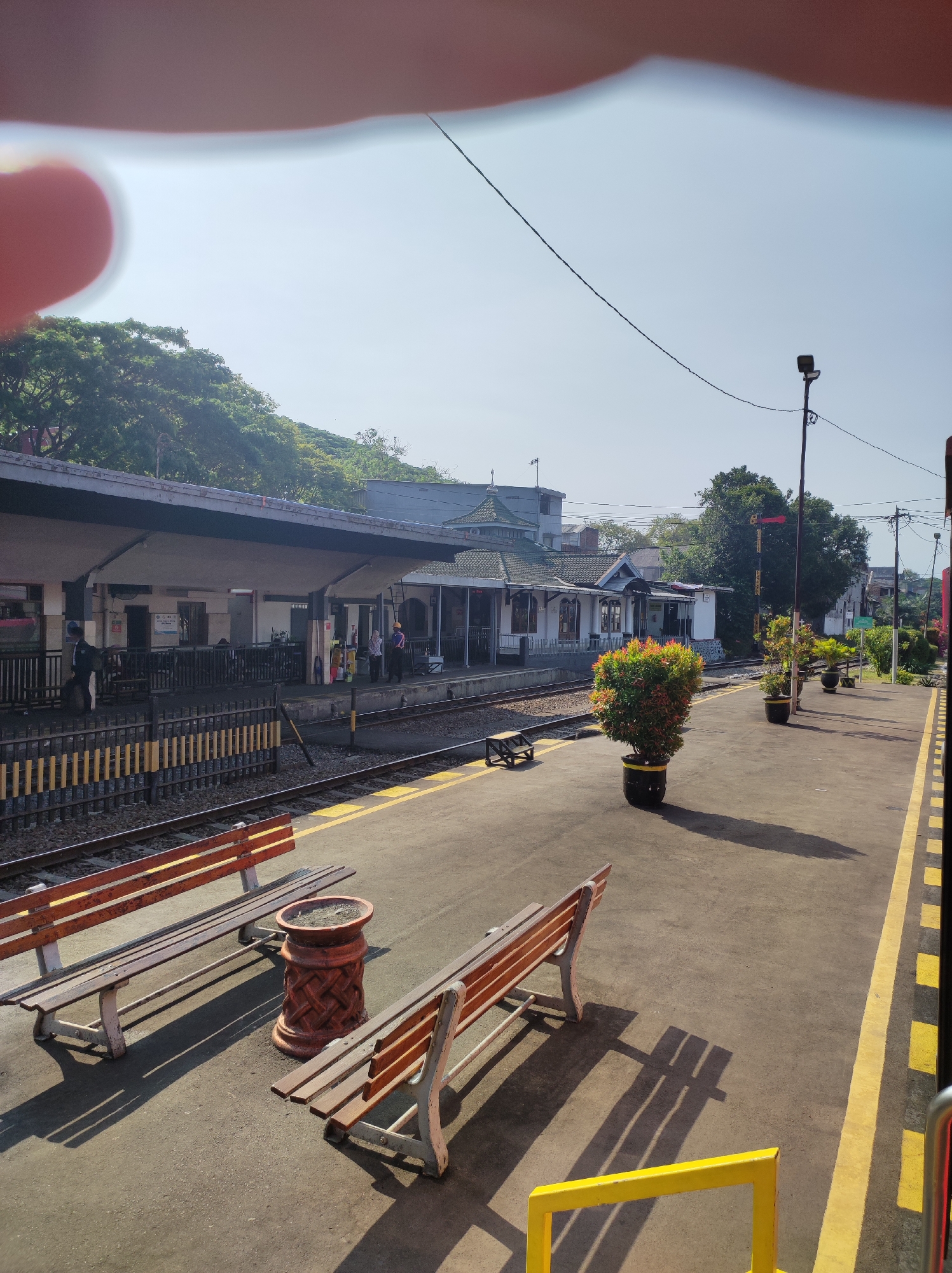 The image captures a sunny day at a train station in Malang, Indonesia. A red-brown wooden bench with a white metal frame is positioned on the platform, casting a long shadow. Adjacent to it is a decorative, reddish-brown planter with an intricate woven pattern. Another similar bench is visible further down the platform.
The train tracks run alongside the platform, separated by a yellow and black striped safety line. Beyond the tracks, a covered station building with a grey roof and white walls is visible. Several potted plants, including a large bushy plant with red and green leaves in a black pot with yellow accents, line the platform. A signal pole with a red signal light is also present.
In the background, lush green trees cover a hillside, suggesting a natural setting around the urban area. The sky is a clear, pale blue, indicating fair weather. A few people are present at the station, including a uniformed individual near the building entrance and others standing on the platform. The overall scene depicts a tranquil and operational train station.