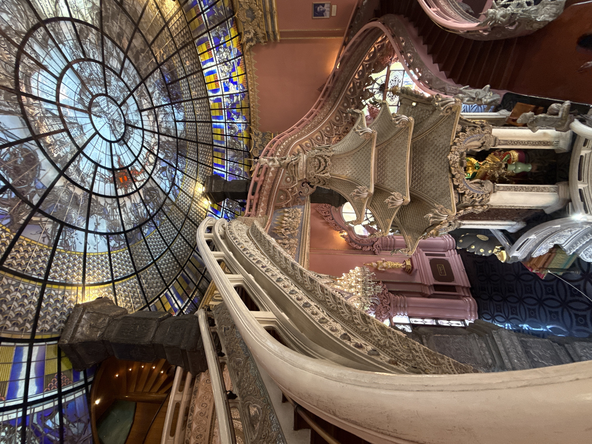 The image captures an ornate interior in the city of Samrong Nuea Subdistrict Municipality, Thailand. A massive stained-glass dome with intricate circular patterns dominates the scene, showcasing a blue and yellow color scheme. Adjacent to the dome is a winding, highly decorated staircase with white railings and detailed carvings in a light pink and beige color palette. The staircase ascends past openings offering glimpses of interior spaces and additional staircases. The environment exhibits a blend of architectural styles, showcasing Thai artistry.