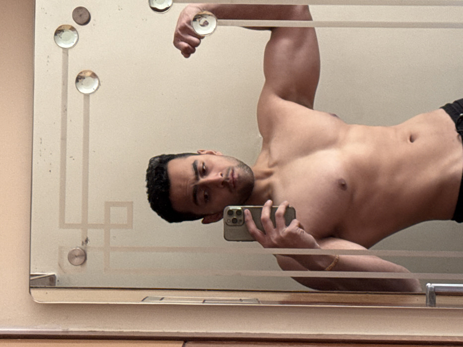 A man is taking a selfie in a mirror. The mirror has decorative glass buttons around the edges. He is shirtless and wearing black shorts. He is holding his phone in his left hand, and his arm is bent at the elbow. His other arm is raised above his head, also bent at the elbow. He has dark hair and a slight beard. His reflection is visible in the mirror.