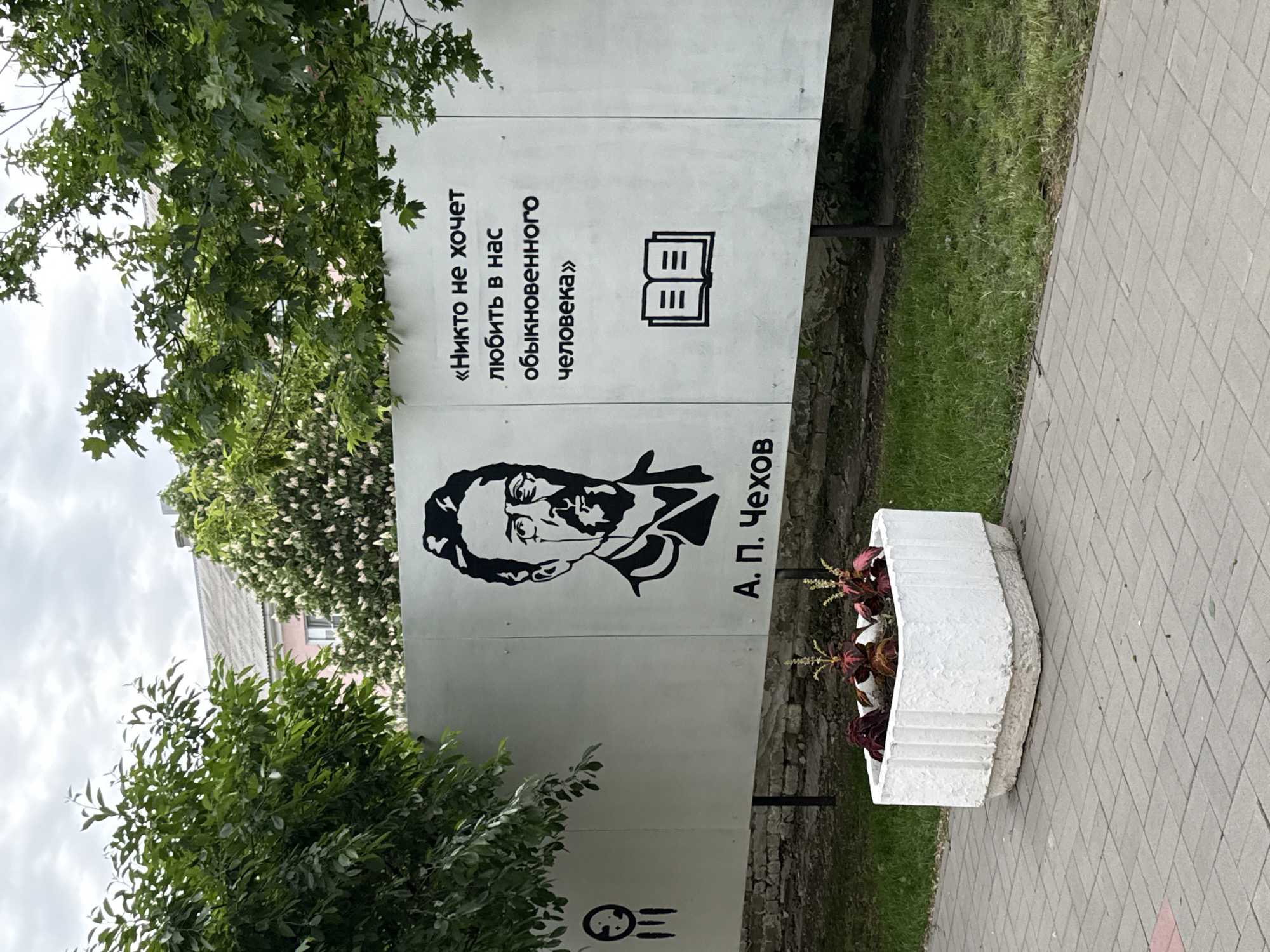 The image captures a street scene in Uryupinsk, Russia. A large, vertical white sign stands prominently on the street. The sign features a black and white portrait of Anton Chekhov, and beside his portrait, the name "A. П. Чехов" is written in Cyrillic. Above the portrait is a quote in Russian and an icon of a book. To the right of the sign is a white, concrete flower planter filled with red and green plants. The sign is surrounded by greenery, including trees. The paving stones of the sidewalk are visible on the right of the sign.