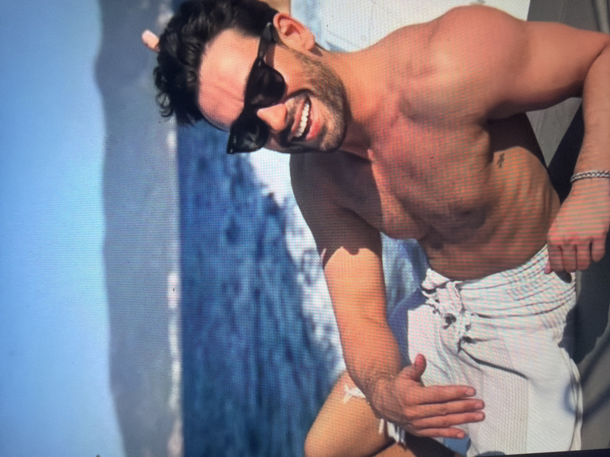 The image is a photograph of a digital screen, exhibiting a visible pixel grid pattern across its surface. It depicts an adult male, likely in his late 20s or 30s, reclining in bright sunlight.
The man is bare-chested, showcasing a muscular physique and tan skin. He wears light-colored, possibly white or light grey, drawstring shorts or swim trunks. His hair is dark and curly, and he has a short beard or stubble. He is wearing dark, rectangular-frame sunglasses. His face is turned towards the viewer, displaying a wide, joyful smile with visible white teeth. A silver-toned chain-link bracelet is present on his left wrist. His right arm is bent with his hand resting near his hip, and his left arm is also bent with his hand resting on his lower abdomen, suggesting a relaxed pose.
He is lying on a light-colored surface, possibly a boat deck or sun lounger. Behind him is a large body of deep blue water, featuring a visible white wake or splash, indicating movement or a watercraft. In the distant background, hazy, dark landmasses (hills or mountains) are visible beneath a clear, light blue sky.
The scene strongly suggests a coastal or large lake environment. There is no visual information within the image itself to indicate the location is the city of Shumen, Bulgaria, which is an inland city and does not feature such a marine landscape.