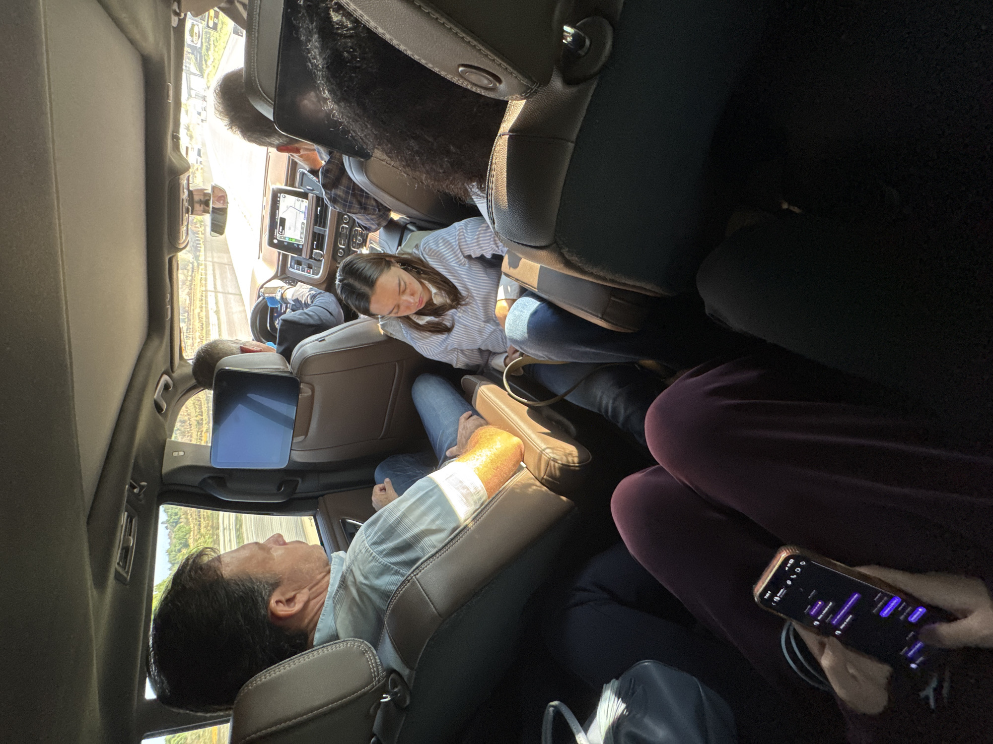 The interior of a vehicle is filled with passengers. An adult male is reclining in the seat with a headrest monitor. Another adult female sits in the middle row, and another adult is sitting in the seat, looking at a phone. The car is presumably moving along a road near the city of San Miguel la Presa, Mexico.