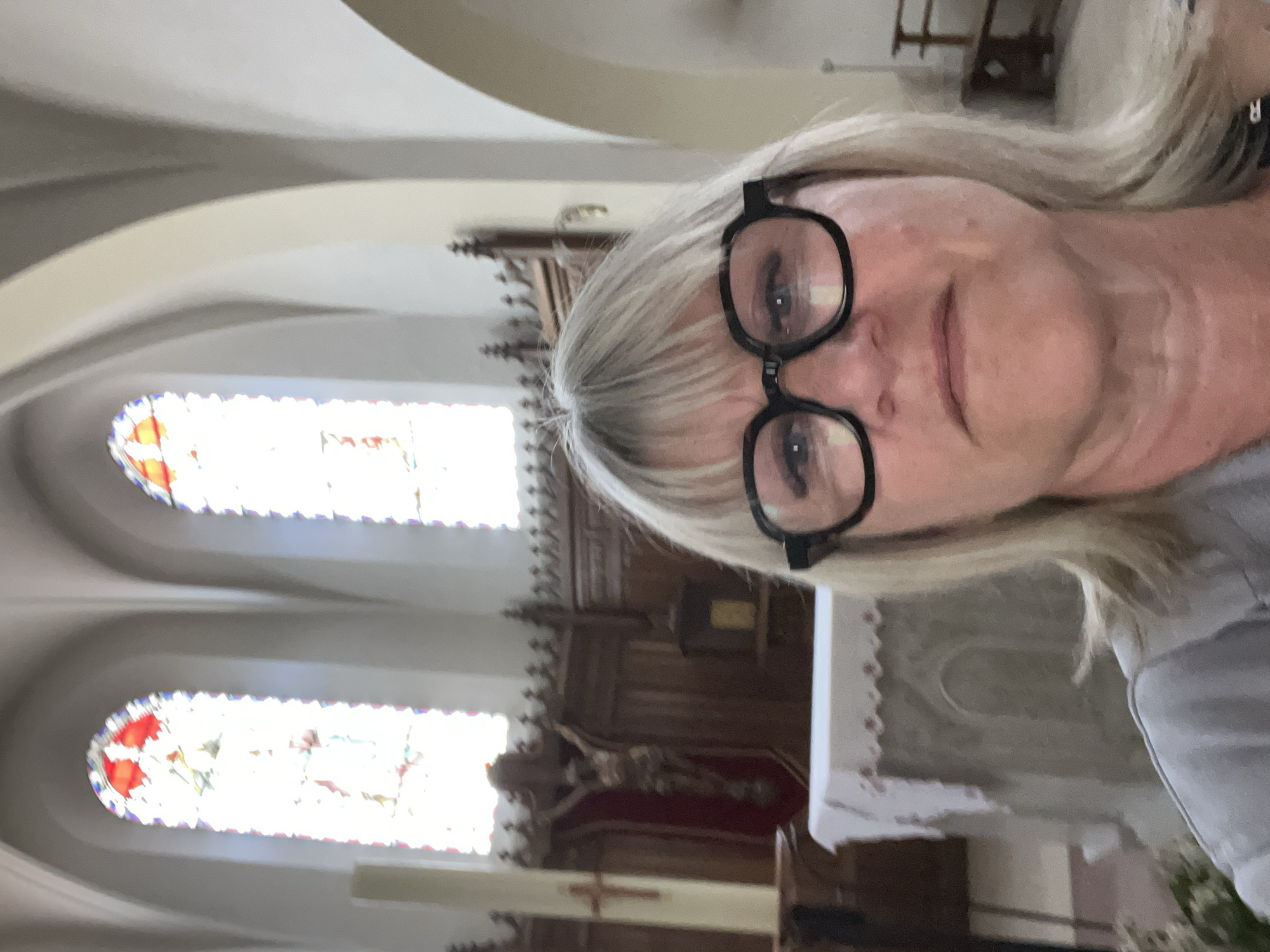 A selfie taken inside the Église Saint-Pierre, located in Chambœuf, France. The image is a medium close-up of a woman with fair skin and blonde hair, wearing black framed eyeglasses. The background reveals the interior of the church, including stained glass windows and an altar. The image is rotated 90 degrees counterclockwise.