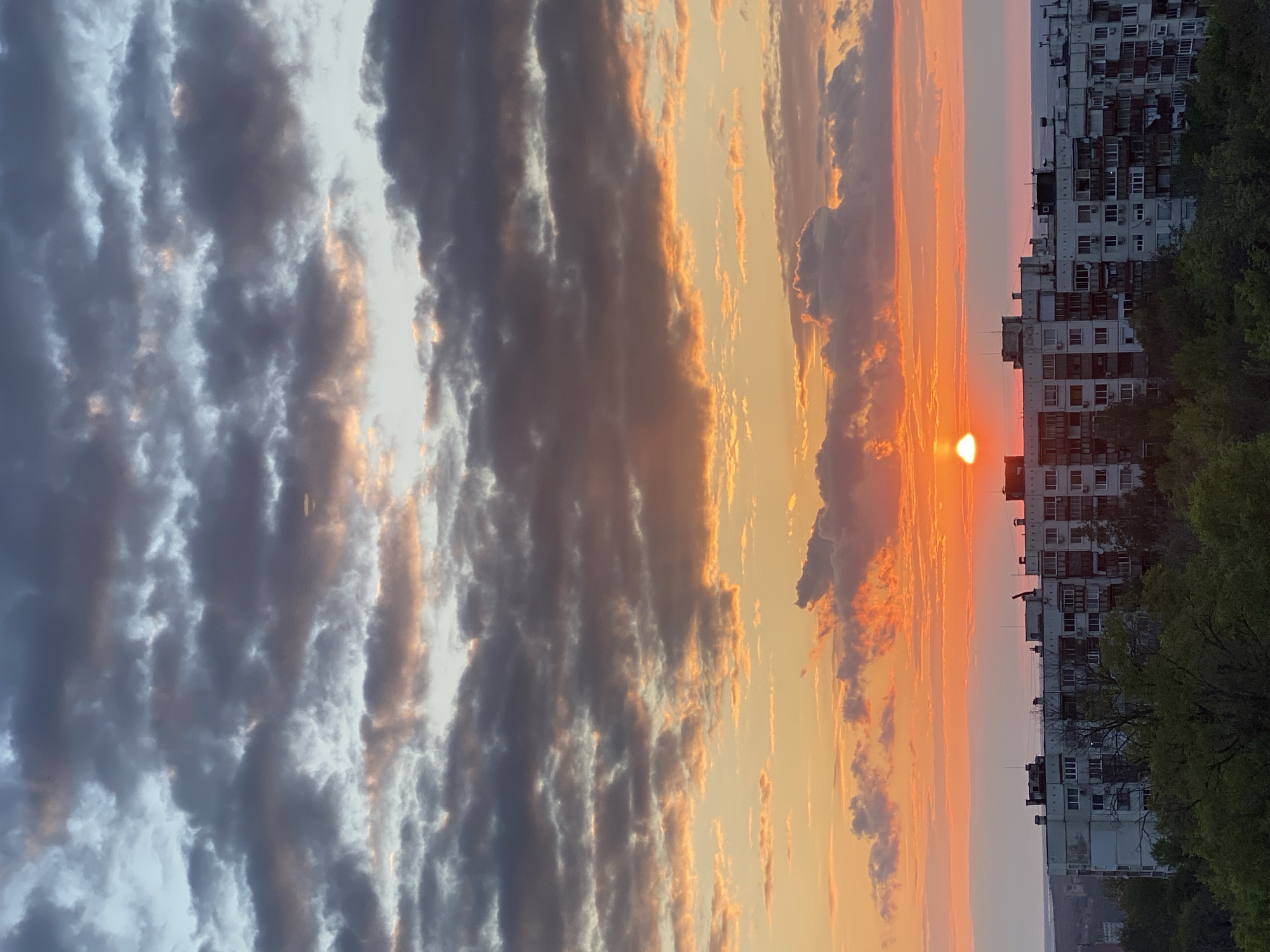 The image shows a sunset in the city of Pleven, Bulgaria. The sun is visible as a bright orange disk just above the buildings, creating an orange glow in the sky. The sky is filled with clouds, with some darker clouds on the left side of the image and lighter, more colorful clouds near the sun. A tall apartment building is located on the right of the image, with green trees partially obscuring it.