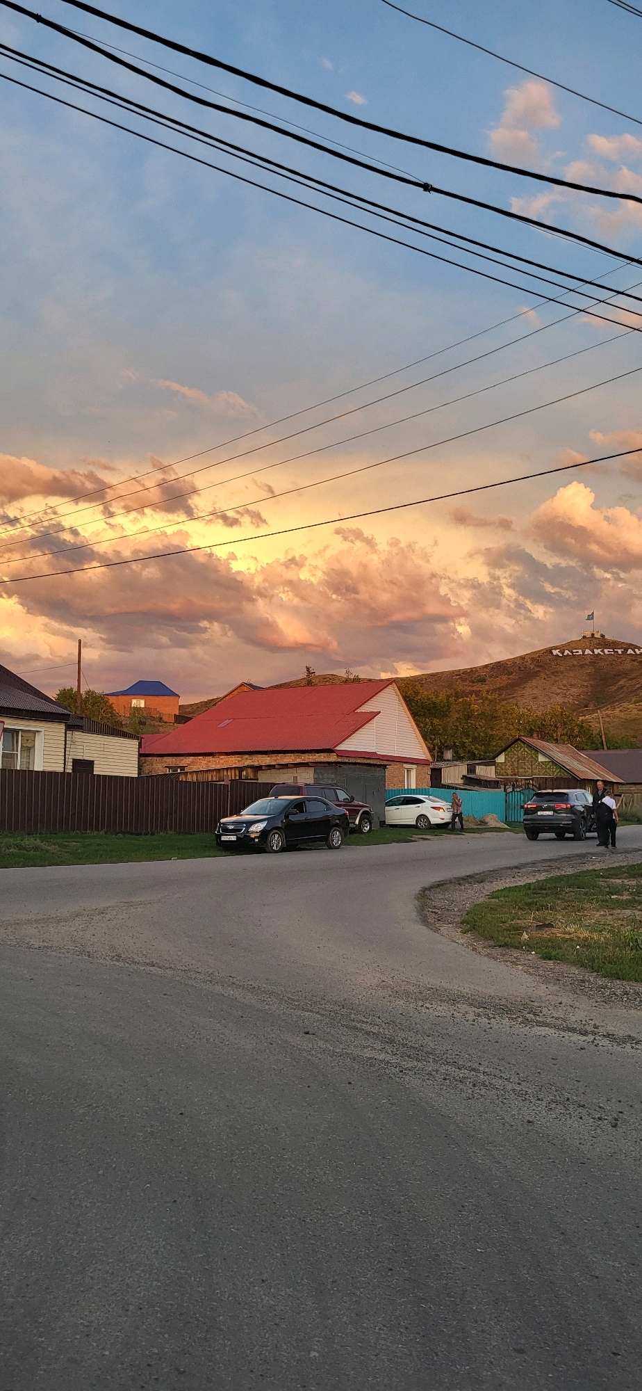 An outdoor, eye-level, vertical, full shot captures a rural street scene at sunset, with a dramatic sky filled with vibrant orange and yellow clouds. The foreground is dominated by an asphalt road that curves to the right. On the left side of the road, several houses with brown fences and red roofs are visible. A black sedan is parked in front of one of the houses. Further down the road, other houses and a light blue fence are present. In the background, a hill rises, and large white letters spelling "ҚАЗАҚСТАН" (Kazakhstan) are displayed on its slope, topped by a flag. Two people, dressed in dark clothing, are standing near a dark SUV on the right side of the street. The overall impression is of a quiet, possibly remote, community bathed in the warm glow of twilight.