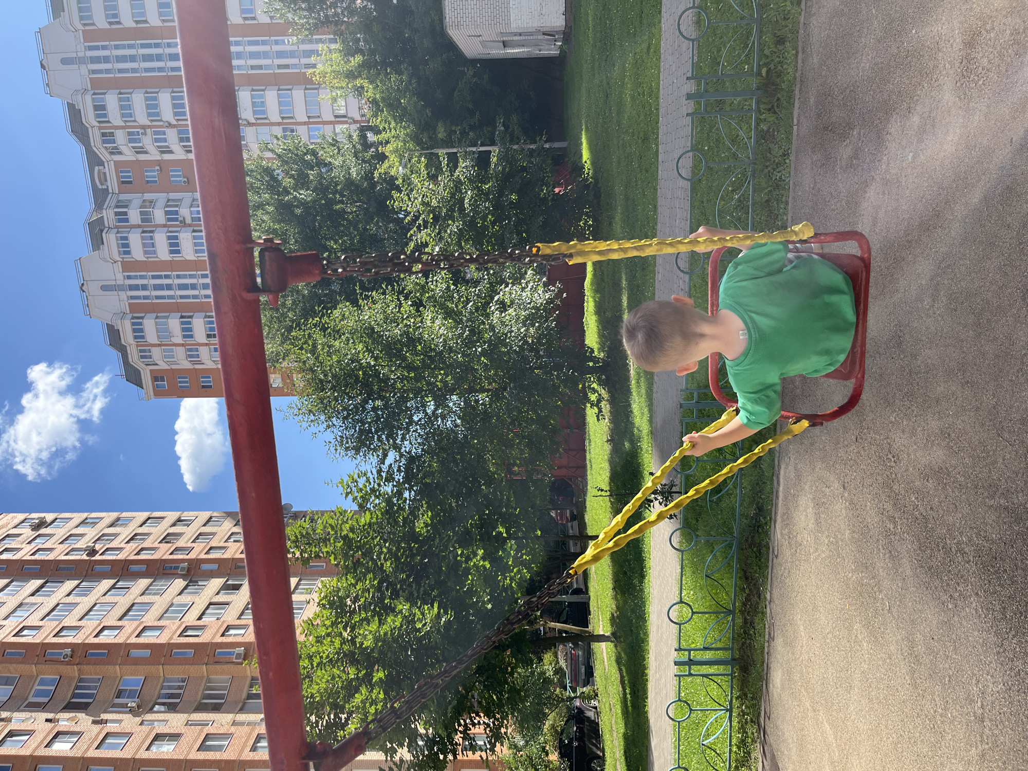 The image captures a young boy, viewed from behind, sitting on a red swing with yellow rope handles. He is wearing a green t-shirt. The swing set is situated on a paved area in what appears to be a residential courtyard. Behind the swing set, a green lawn is visible, bordered by a decorative metal fence. Beyond the lawn, trees provide a leafy backdrop. In the distance, towering apartment buildings, characteristic of urban environments, dominate the skyline. The buildings are modern in design, with numerous windows and balconies. The sky above is a clear blue, with a few fluffy white clouds. This scene is located in the city of Balashikha, Russia.