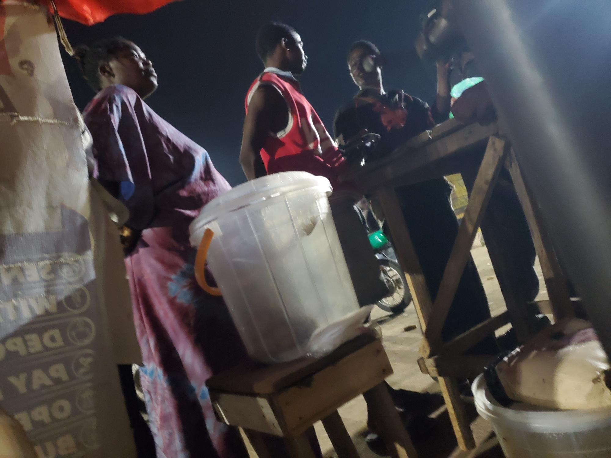The image depicts a nighttime scene in Gwong, Nigeria, focusing on a group of people gathered around a makeshift stall. The stall appears to be constructed from wood. In the foreground, a large, transparent plastic container with an orange handle sits on a wooden stand. To the left, a woman is partially visible, wearing a patterned robe. To the right, two men are standing. One man, wearing a red vest over a dark shirt, is looking to his left. Another man, wearing a dark shirt with a design, is also looking towards the left. In the background, a motorcycle is visible. The overall lighting suggests a low-light environment, possibly an outdoor market or gathering.