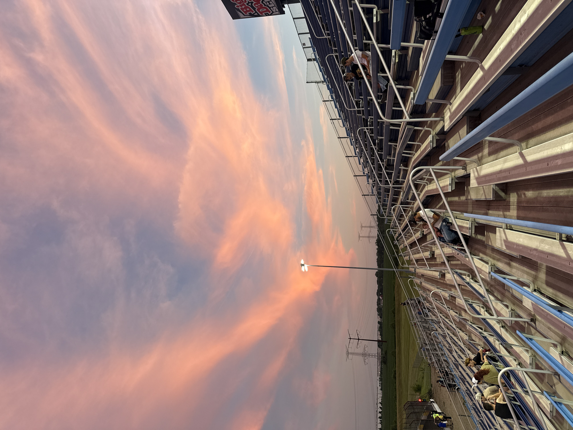 This photograph depicts a sunset scene at a stadium, likely in Joliet, Illinois, given the prominent presence of large electrical transmission towers in the background, which are characteristic of power infrastructure in the region. The sky is dramatically lit with streaks of pink and orange clouds, indicative of a vibrant sunset. In the foreground, tiered bleacher seating, typically found in sporting venues or outdoor event spaces, is visible. Some individuals are seated on the bleachers, observing the sky or engaging in conversation. The stadium lighting is on, casting shadows across the seating area. The overall environment suggests an outdoor event or a gathering taking place at dusk.