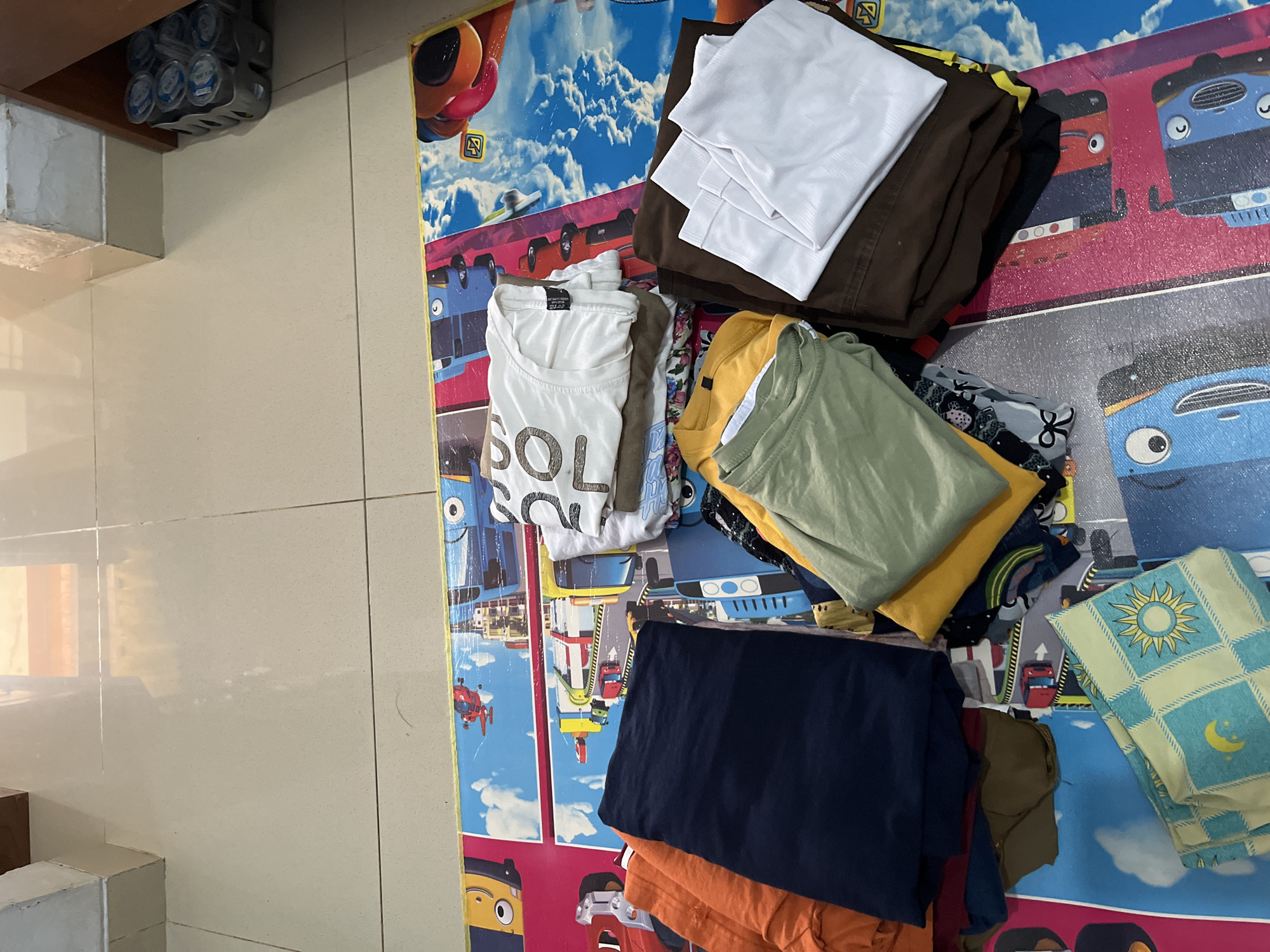The image displays a pile of folded clothes on a colorful children's playmat. The playmat features cartoon vehicles and a sky with clouds. The clothes are a mix of t-shirts and pants in various colors, including orange, navy blue, white, olive green, yellow, and brown. Some of the t-shirts are plain, while others have graphic prints. The clothes appear to be neatly folded. In the background, part of a tiled floor and a wooden cabinet can be seen. A grey plastic holder containing several plastic cups is visible on top of the cabinet. The scene is indoors, likely in a residential setting in Glagah Kulon, Indonesia.