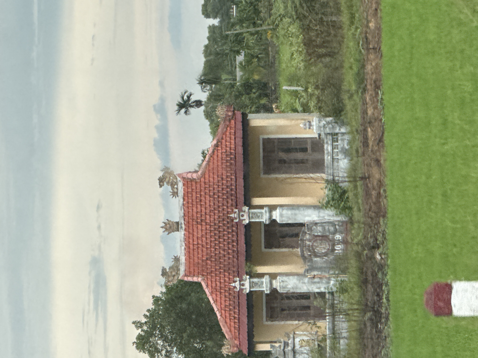 This is a medium-shot photograph of a small, yellow building with a red tiled roof. The building appears to be a temple or shrine, as indicated by decorative statues of mythical creatures on the roof ridge and ornate pillars flanking the entrance. The building has two prominent windows on its facade, and a decorative metal gate in front. The number "1079" is visible on a stone structure to the right of the gate. The building is situated in a rural setting, with lush green fields in the foreground and a dense backdrop of trees and vegetation. The sky is overcast with a few wispy clouds. A red and white marker post is visible in the lower left corner of the frame. The image is taken from a slightly elevated position, likely from a vehicle. The location is Hiếu Giang Commune, Vietnam.