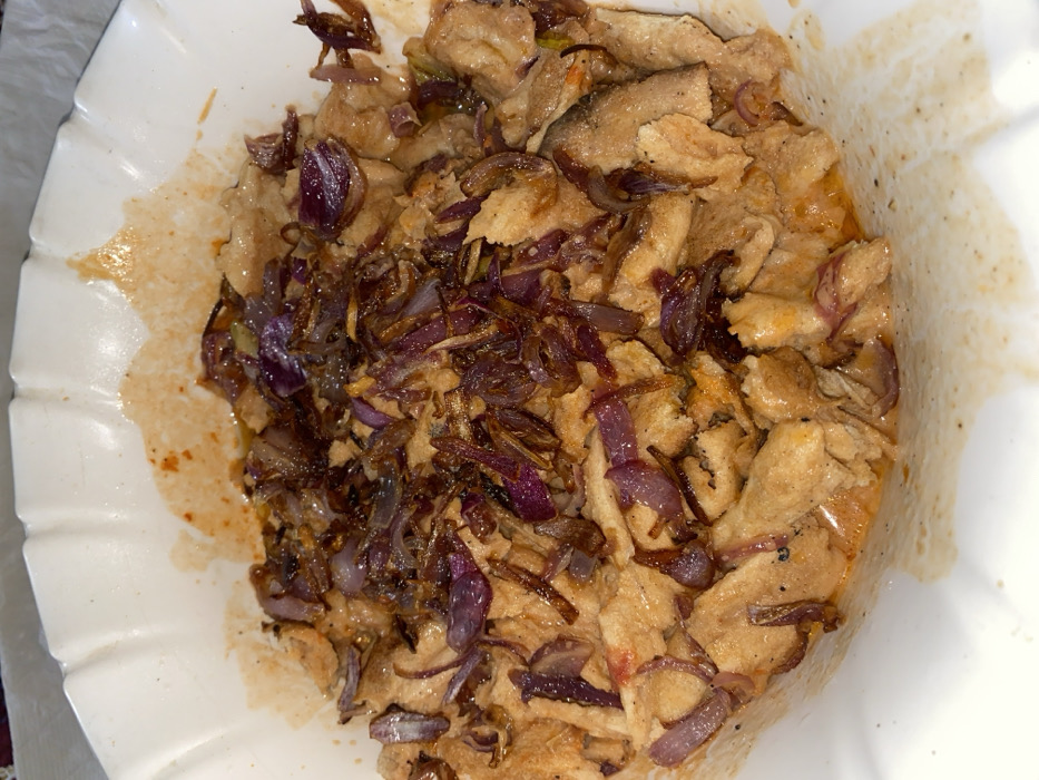 The image displays a close-up, overhead view of a hearty meal served in a white, scalloped-edge dish or bowl. The primary components of the dish are a base of light brown, irregularly shaped pieces of food, heavily topped with generous amounts of caramelized red onions.
The main body of the food consists of numerous chunks that appear to be cooked meat, possibly chicken or turkey, or a similar textured protein. These pieces are irregular in shape and size, with a somewhat rough, shredded, or torn texture, and are a muted light brown or beige color. They seem to be soaking in or coated with a light yellowish-orange sauce or liquid, which is visible at the bottom of the dish and glistening slightly. There are also hints of a thin, light brown layer underneath, possibly a piece of flatbread or similar bread product, which appears to line the dish and absorb some of the liquid.
Dominating the top layer of the dish is a substantial quantity of thinly sliced red onions that have been deeply sautéed or caramelized. Their colors vary from a deep purplish-red to dark brown, suggesting a rich, sweet, and savory flavor. Some onion pieces appear softer and more translucent, while others look more crisped. These onions are scattered liberally over the main food pieces, almost completely covering them in many areas.
The white serving dish has a distinct ruffled or scalloped rim. The bottom and sides of the dish show some residue of the yellowish-orange sauce and spice flecks, indicating the saucy nature of the meal. The overall presentation suggests a home-cooked, robust dish, rich in flavor and texture, possibly a type of stew or meat dish served over bread. No people or text are visible in the image.