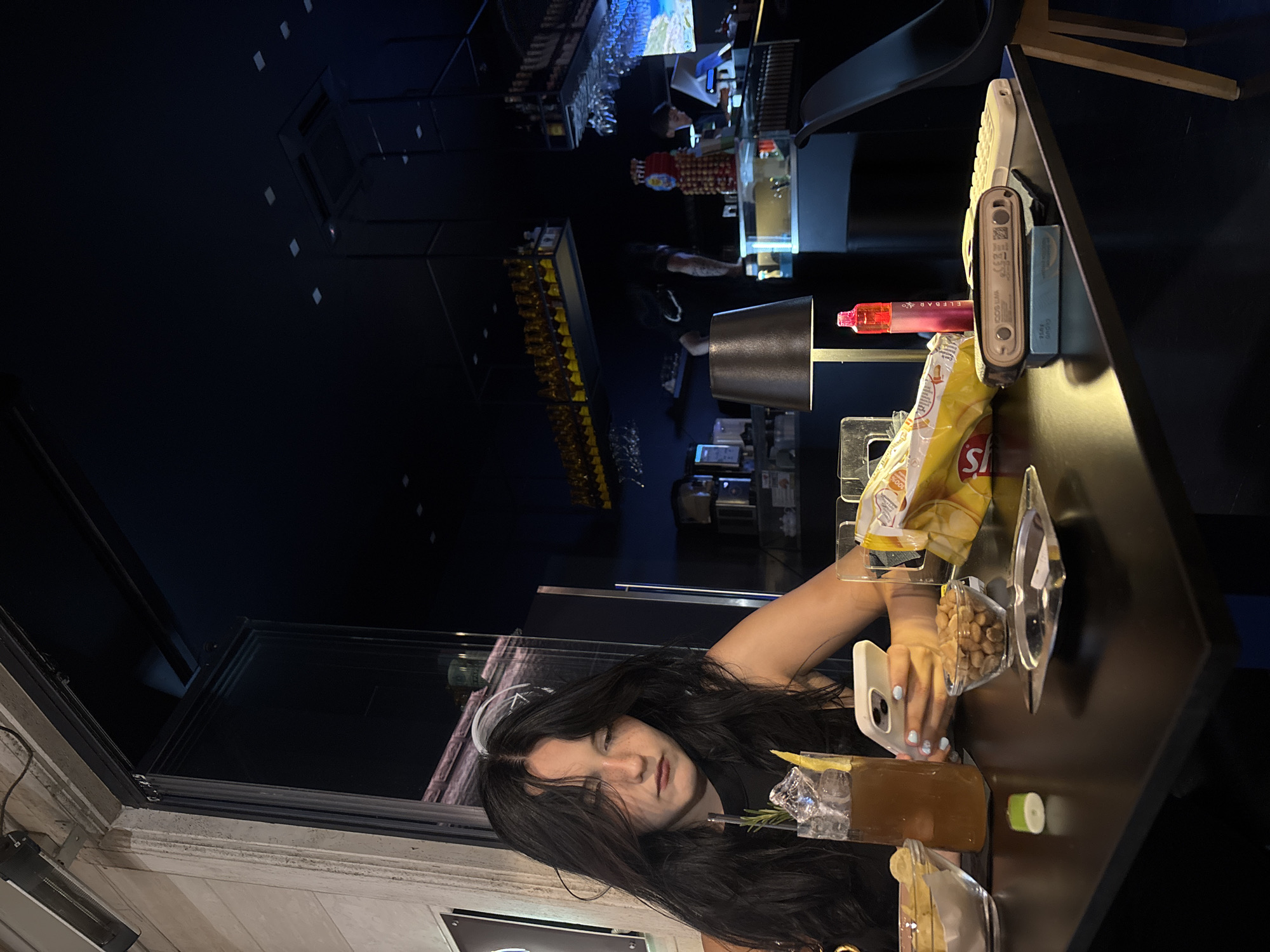 A young woman with long, dark hair is sitting at a black table in what appears to be a bar or lounge. She is looking down at her phone, which she is holding in her right hand. In front of her are a glass of iced tea with a garnish, a bag of chips, a bowl of nuts, and an ashtray with a cigarette butt. To her right, there is a pink vape pen and an electronic cigarette device on a small blue box. The background is dark, with shelves of bottles and bar equipment visible. The overall impression is of a casual, nighttime setting. The specific location in the city of San Benedetto del Tronto, Italy, is not discernible from the image itself.