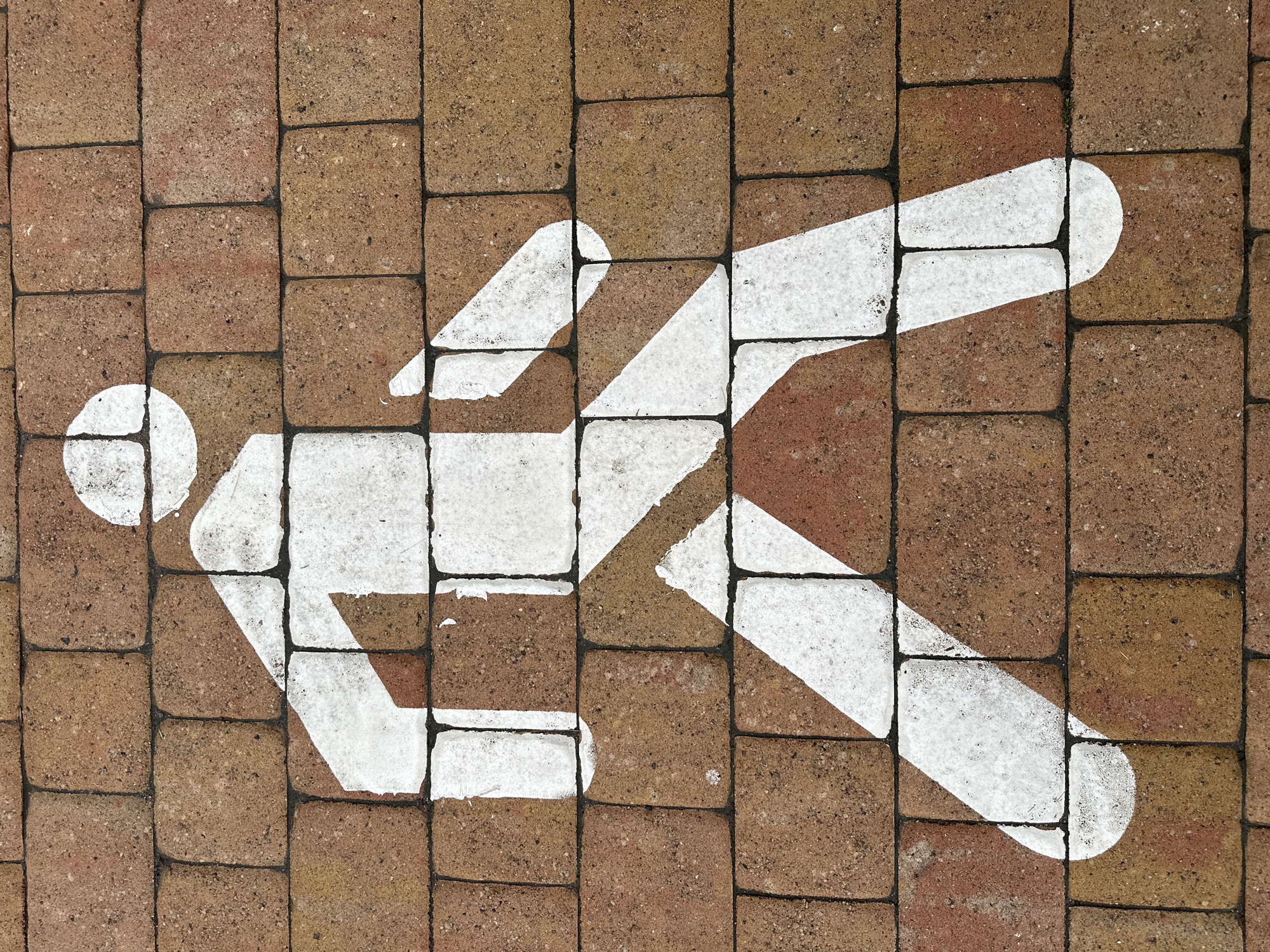 A white pedestrian symbol is painted on a brick walkway in Minsk, Belarus. The symbol depicts a stick figure in mid-stride, with one leg forward and the other back. The figure is simplified, with a circular head, a rectangular torso, and two arms and legs. The brick walkway is made of rectangular bricks laid in a running bond pattern. The bricks are reddish-brown in color and have a rough texture. The symbol is partially worn, with some areas of the paint chipped away. The image is taken from a top-down perspective, looking directly at the symbol on the walkway.