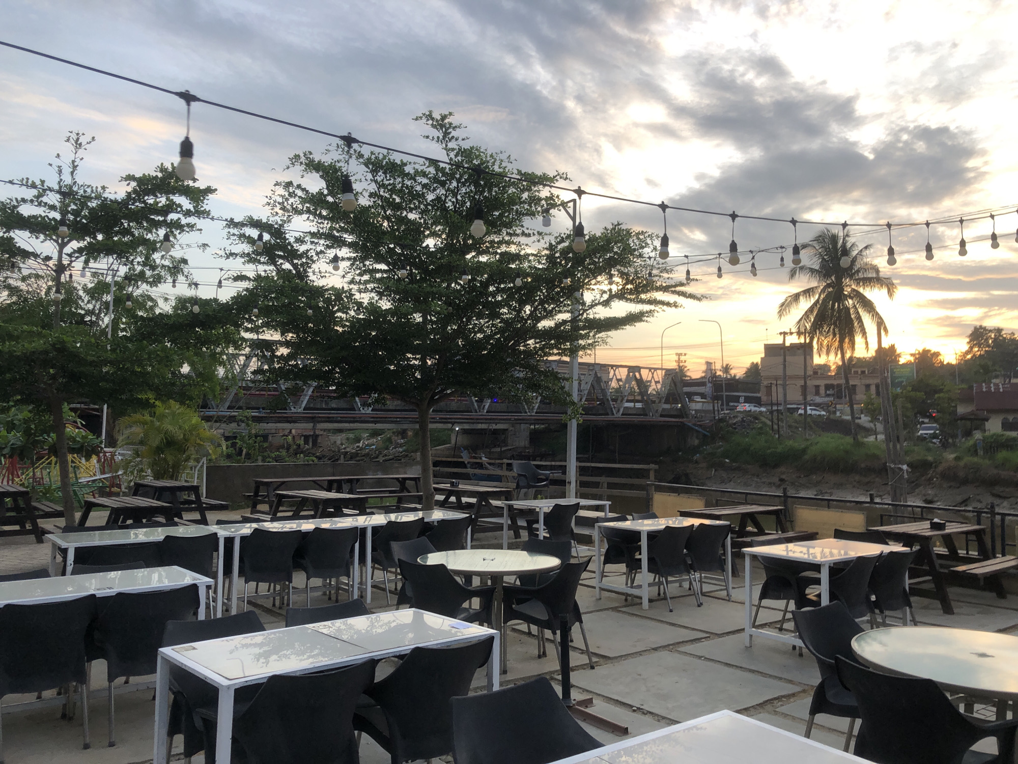 The image depicts an outdoor seating area of a restaurant or cafe, possibly located near the Krueng Pasee river in Idi Rayeuk, Indonesia. The setting appears to be during sunset, with a cloudy sky illuminated by the warm hues of the setting sun. The area is furnished with numerous tables and chairs, predominantly black plastic chairs around white-topped tables, and several picnic-style wooden tables. String lights are suspended overhead, adding to the ambiance. In the background, a bridge structure is visible, along with buildings and trees, including a prominent palm tree. The ground is paved with concrete slabs. There are no people visible in the frame.