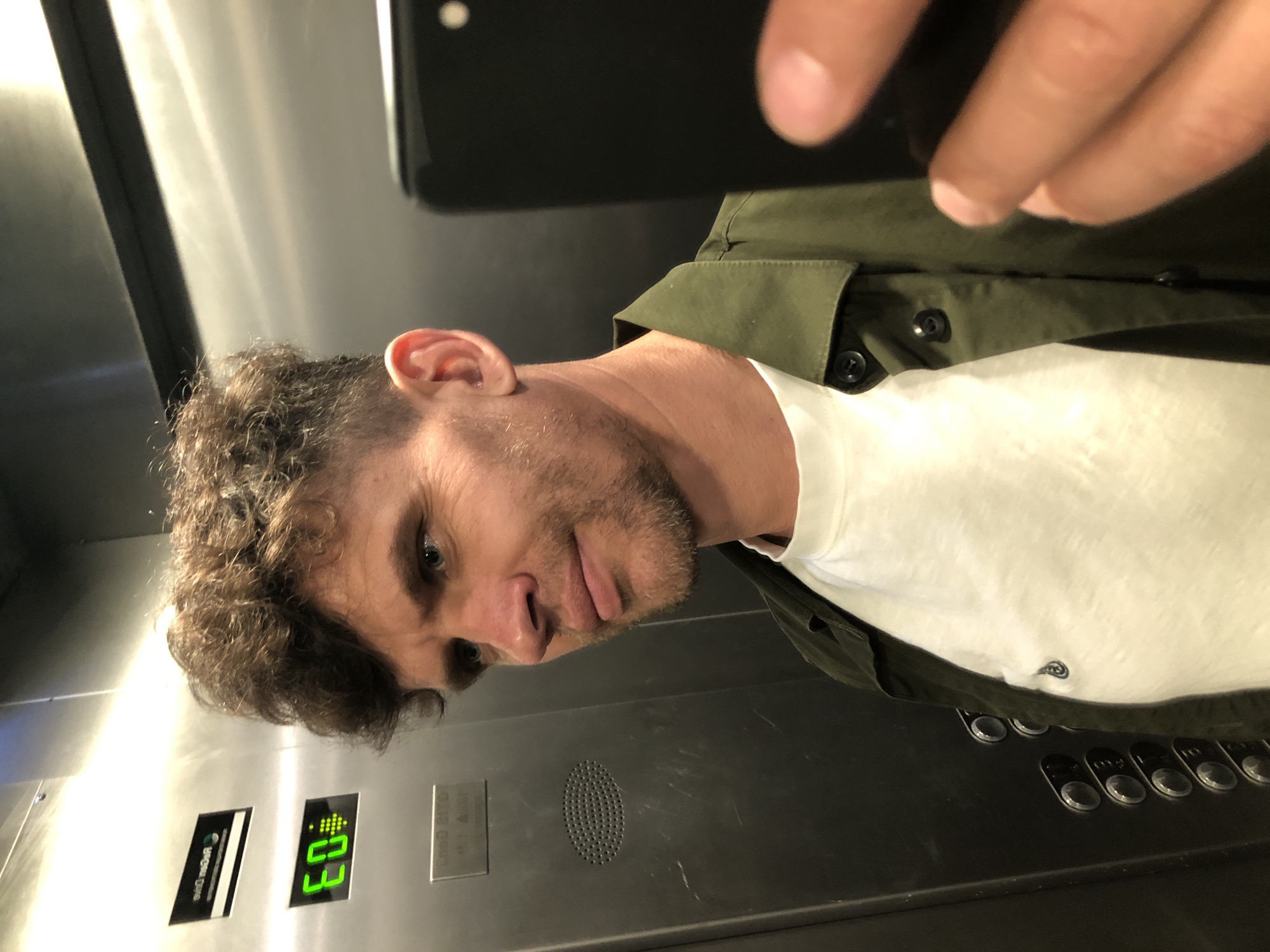 A man with curly brown hair and a beard takes a selfie in an elevator. He is wearing a white t-shirt and a green jacket. The elevator walls are stainless steel, and an illuminated display shows the number "60." Other elevator buttons are visible. The man appears to be in Saint Petersburg, Russia, based on an inscription on a plaque inside the elevator that reads "OTIS."